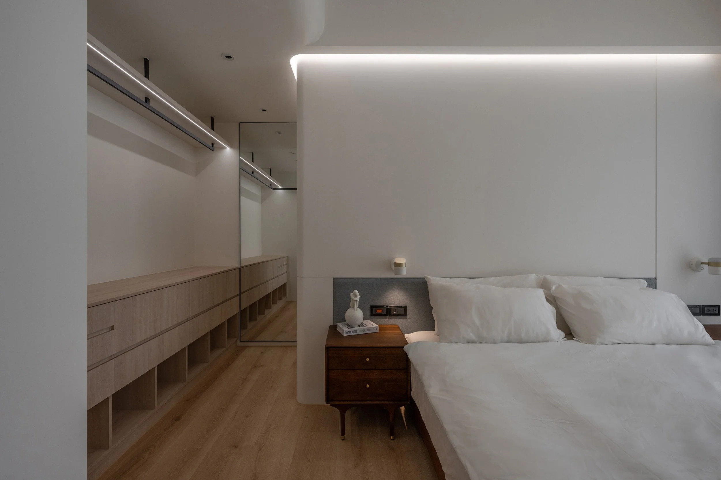 Minimalist bedroom with white walls, a bed with white bedding, a wooden nightstand, a decorative white vase, a large wall mirror, and built-in wooden storage along the left wall, illuminated by modern ceiling lights.