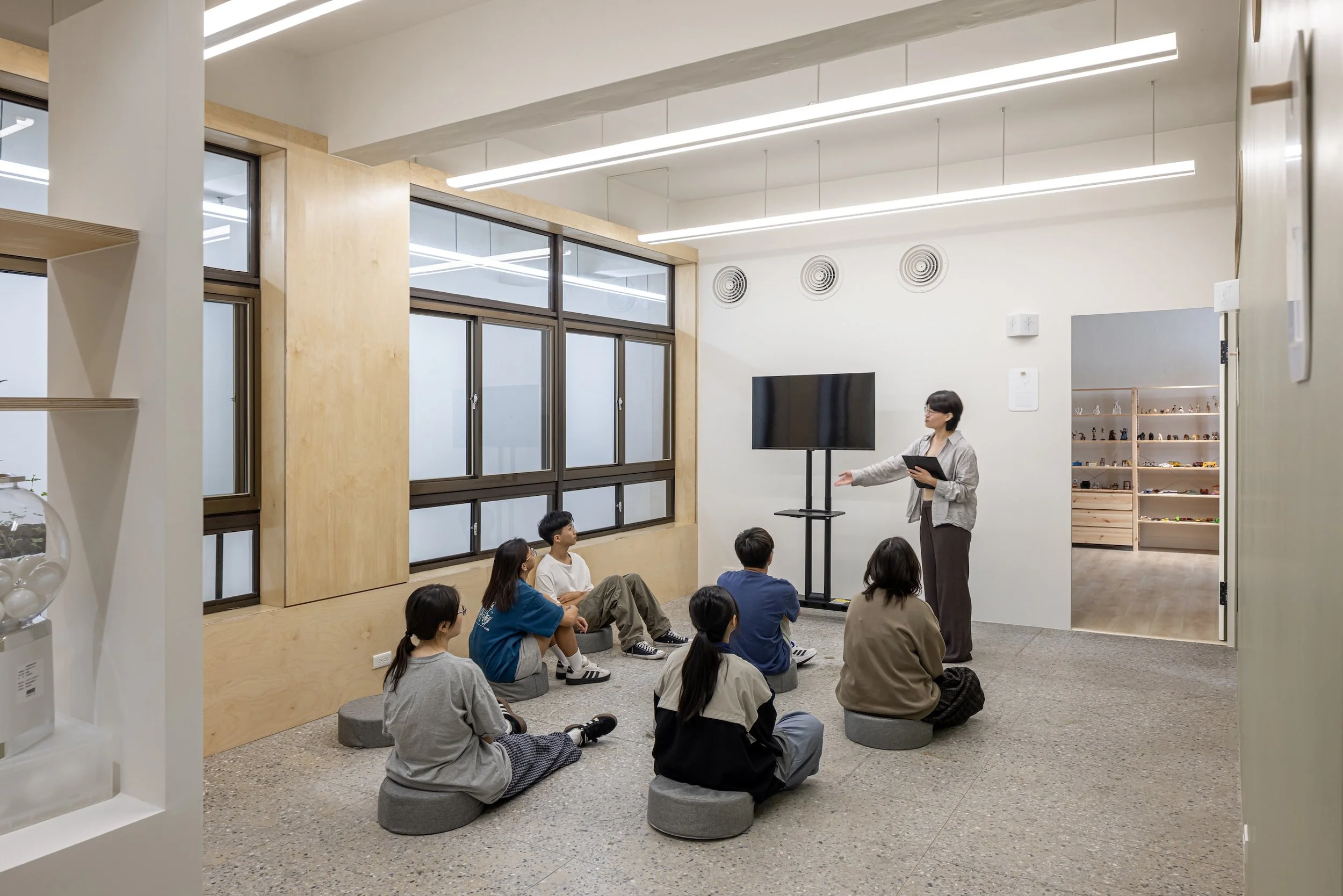 An indoor classroom or workshop setting with a woman instructor standing in front of a group of seated students, some cross-legged on the floor, during a presentation or lesson.