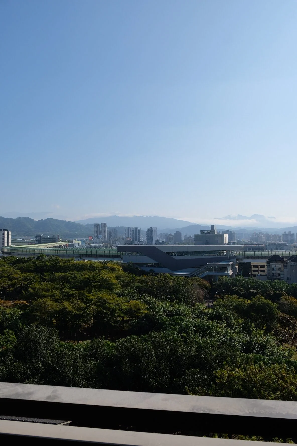 City skyline with modern buildings, mountains in the background, and a green park with trees in the foreground under a clear blue sky.