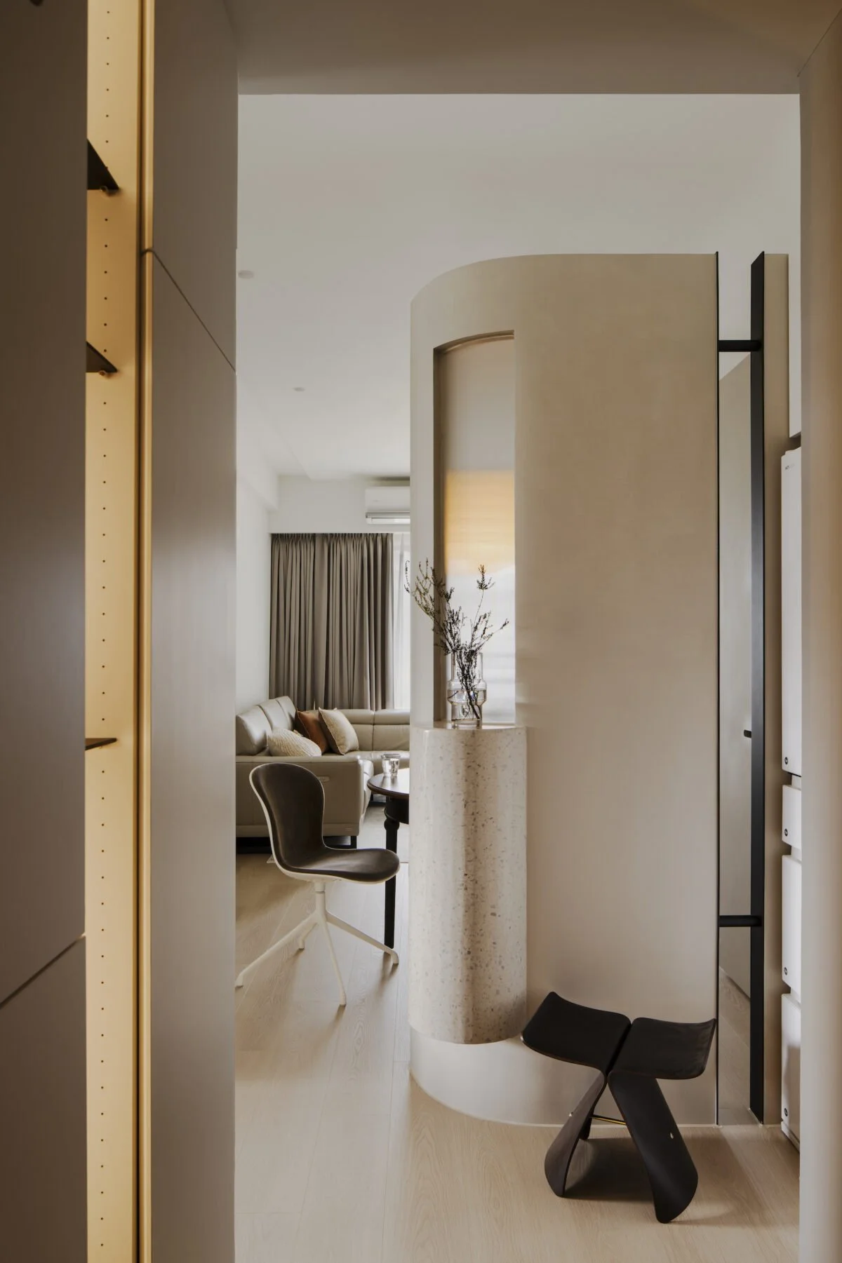 Modern living room with beige walls, a curved partition, a black chair, a beige sectional sofa with cushions, and large windows with beige curtains.