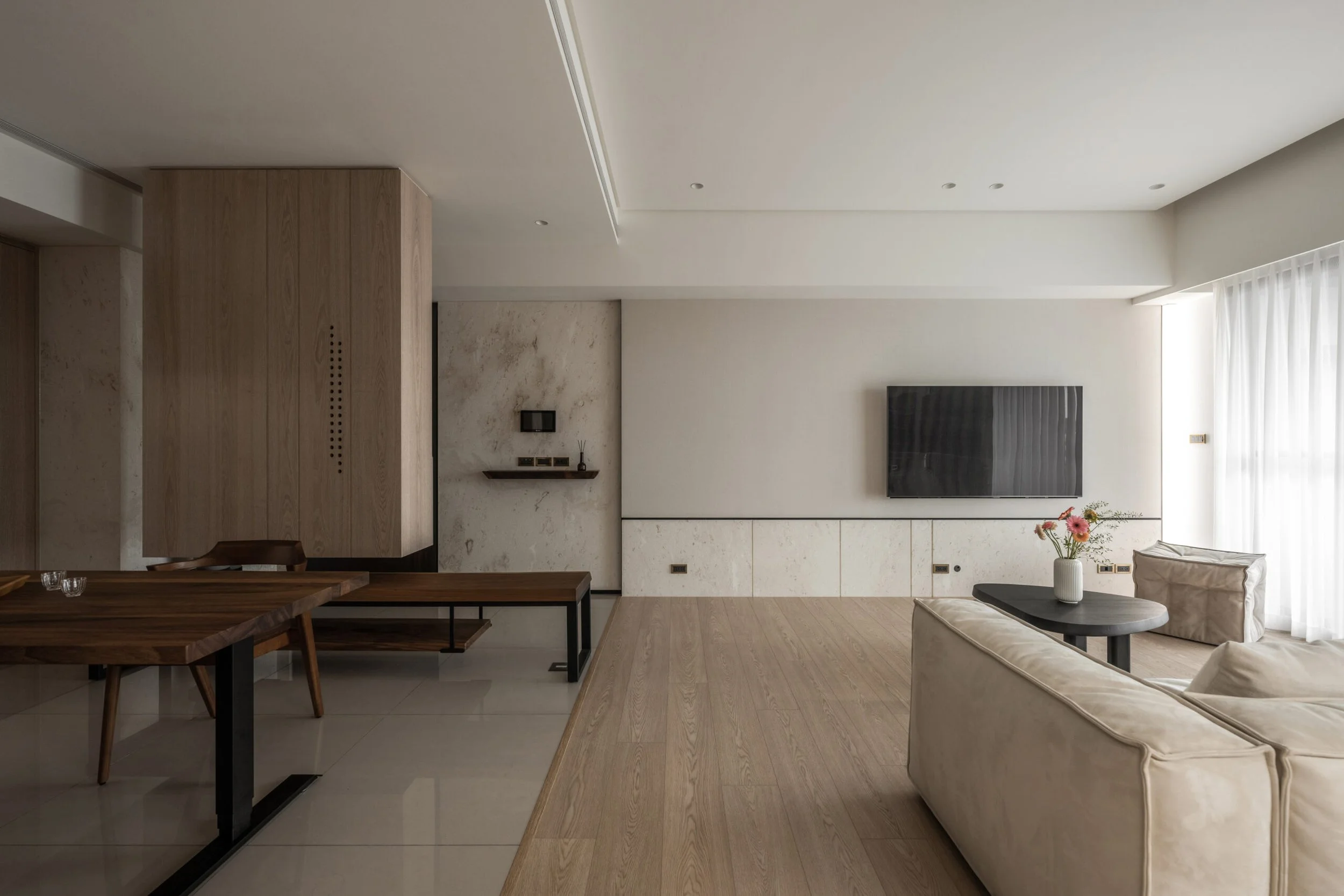 Modern living room with a large wall-mounted television, a beige sofa, a round coffee table with a white vase of flowers, a wooden dining table with chairs, and light-colored curtains on a floor-to-ceiling window.