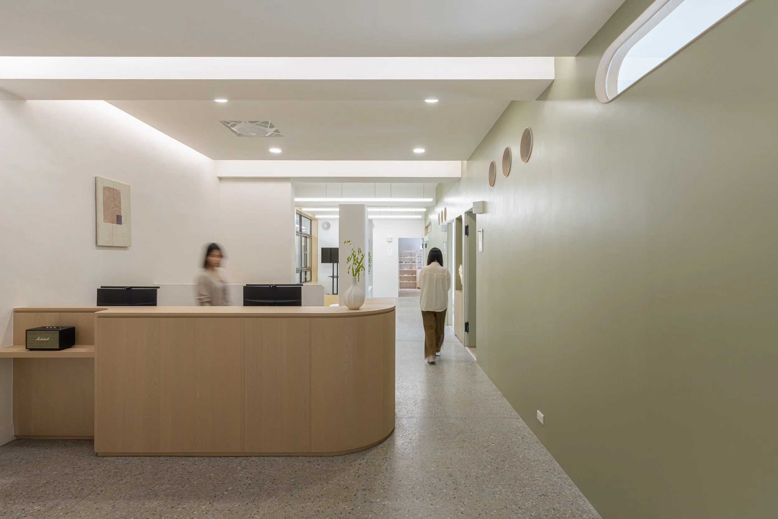 Interior view of a modern, minimalist reception or lobby area with white and light green walls, a curved wooden reception desk with a small amplifier on top, LED ceiling lights, and three people walking or standing in the space. There is artwork on the wall, a large vase with a plant, and windows in the background.