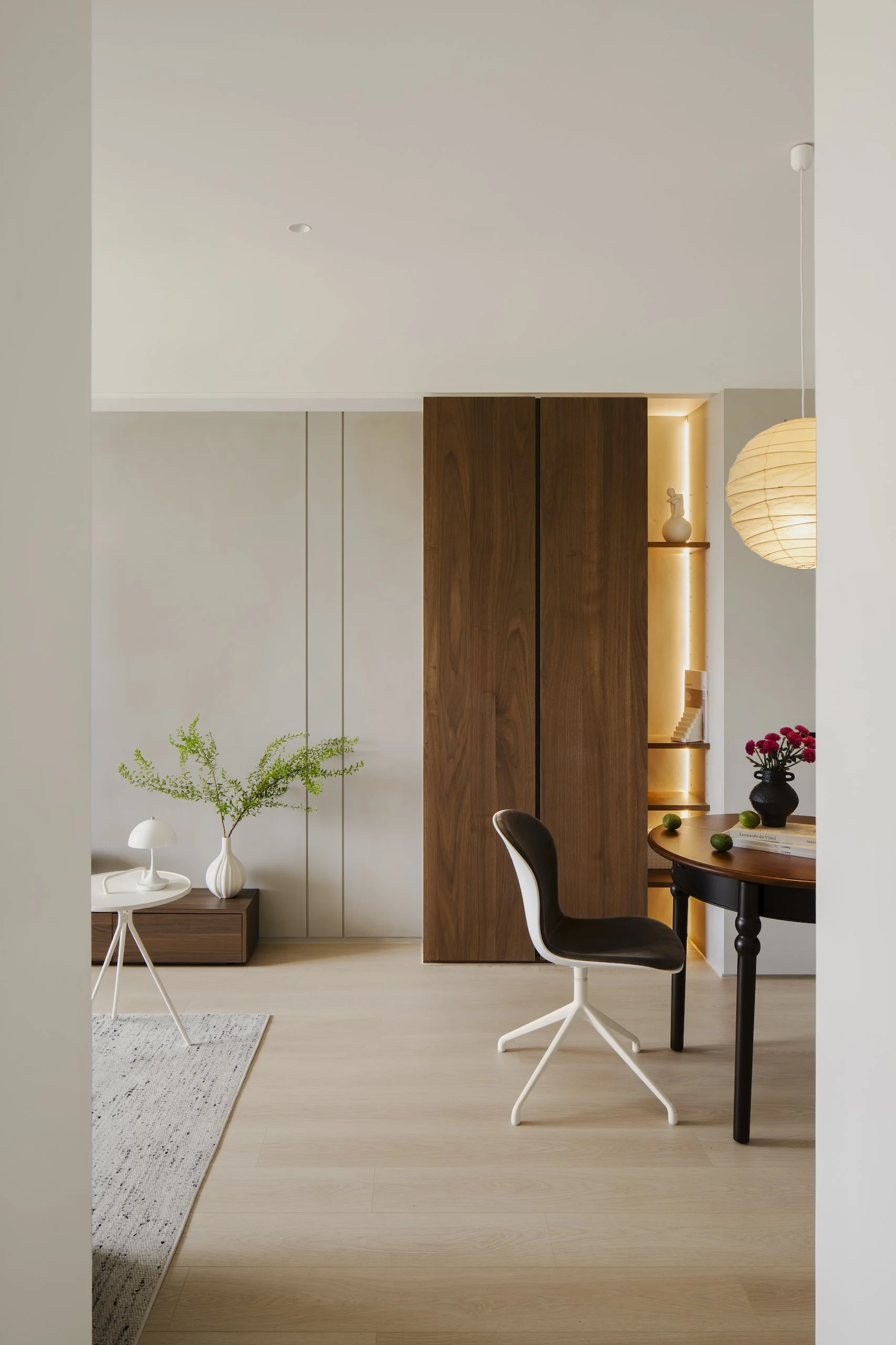 Minimalist modern interior with light-colored wooden flooring, white walls, a dark wood panel, a small side table with a white lamp and a green plant, a round black table with a black chair, decorative items including a black vase with red flowers, a paper lantern pendant light, and a small shelf with a white vase and sculpture.