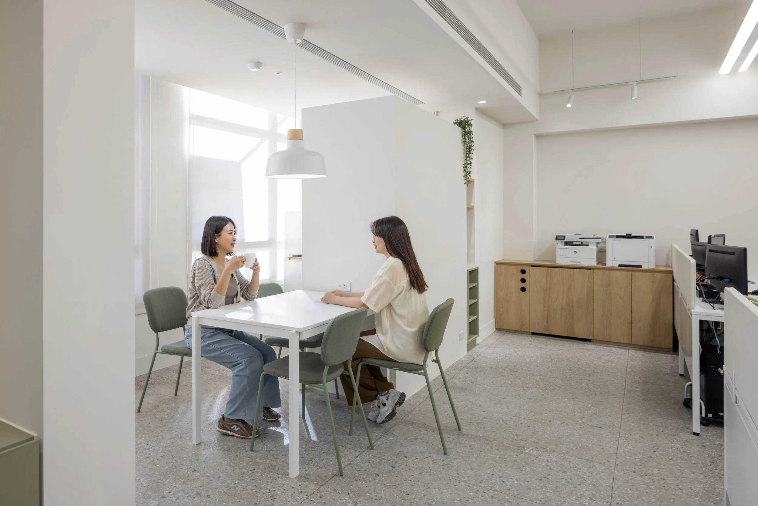 Two women sitting at a white table in an office, engaging in conversation, with a minimalist white interior, windows, filing cabinet, printers, and computer monitors in the background.