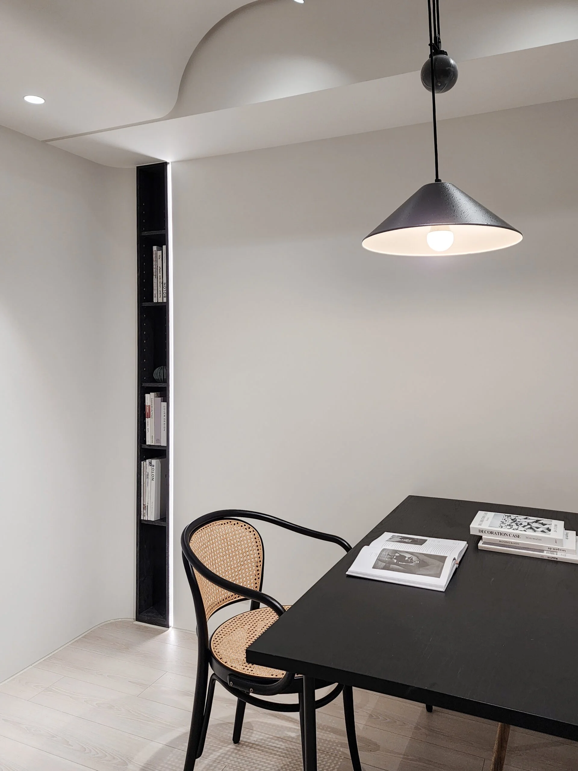 A modern minimalist dining or work area with a black table, two black chairs with rattan woven seats, and decor books on the table. There is a black vertical shelf built into the corner of the room holding books. A black pendant light hangs from the ceiling, and the room has white walls and light wood flooring.