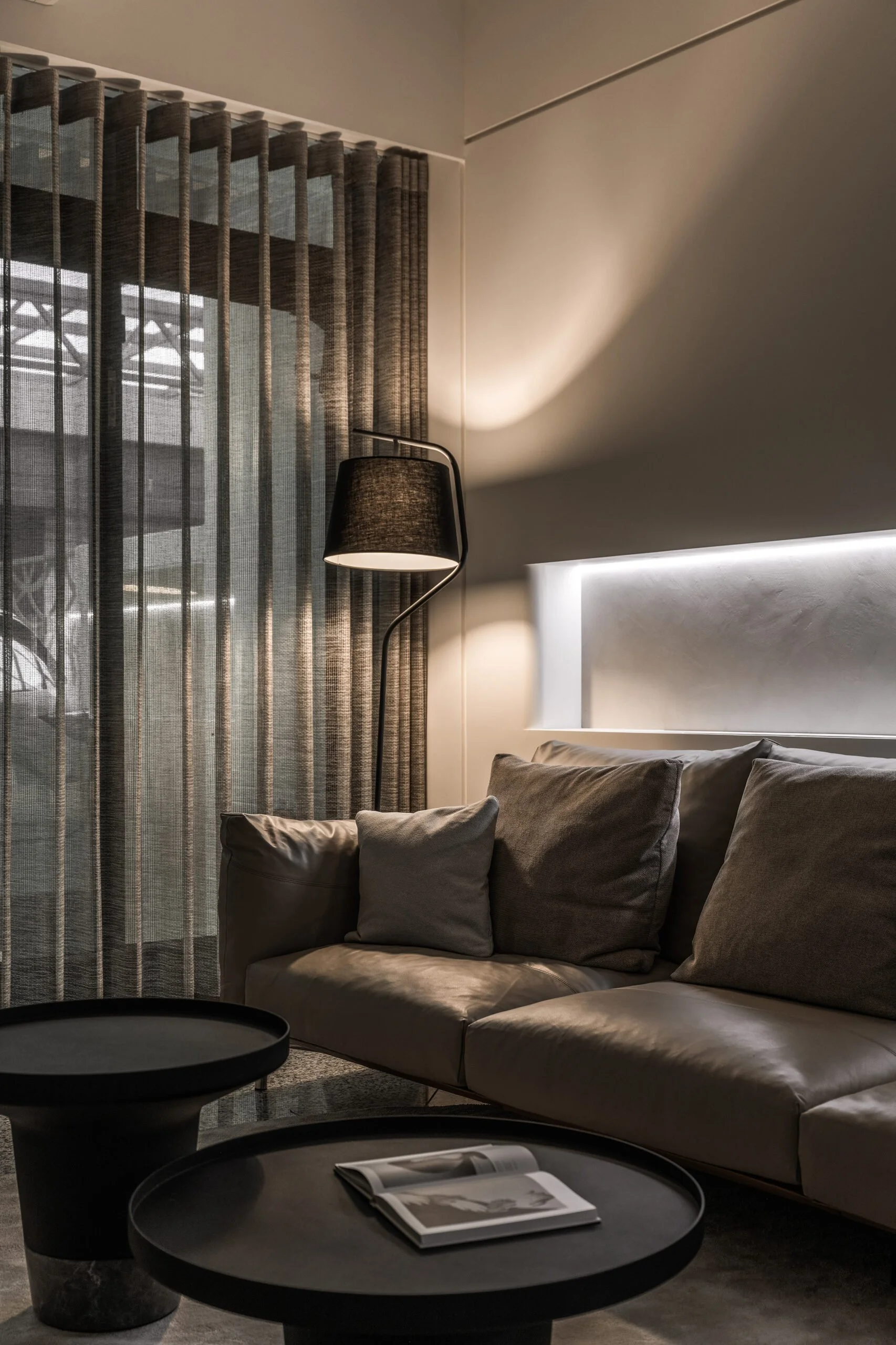 A living room scene with a beige sofa, several gray cushions, a black floor lamp, a window with elongated curtains, a black coffee table with a magazine, and abstract lighting on the wall.