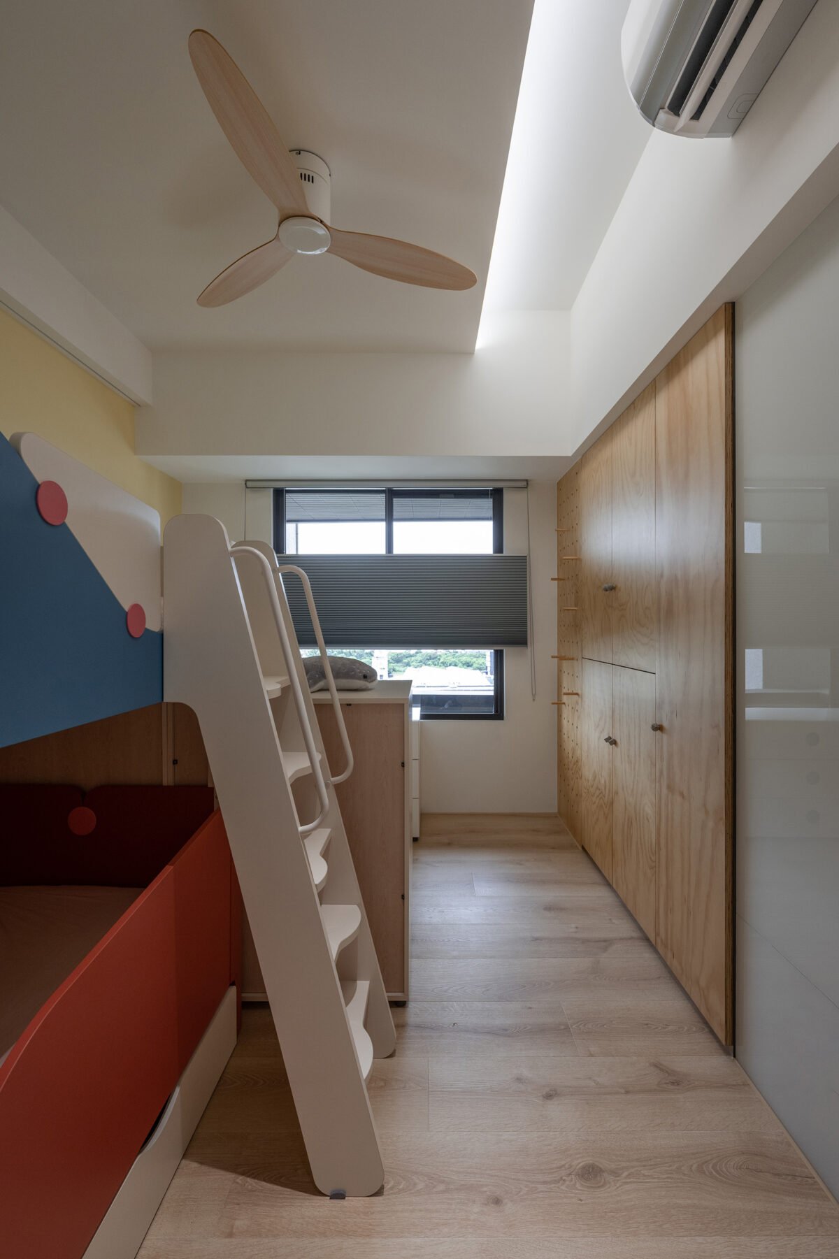 Interior of a modern room with a ceiling fan, air conditioning unit, large window with blinds, wooden cabinet, and a bunk bed with a desk underneath.