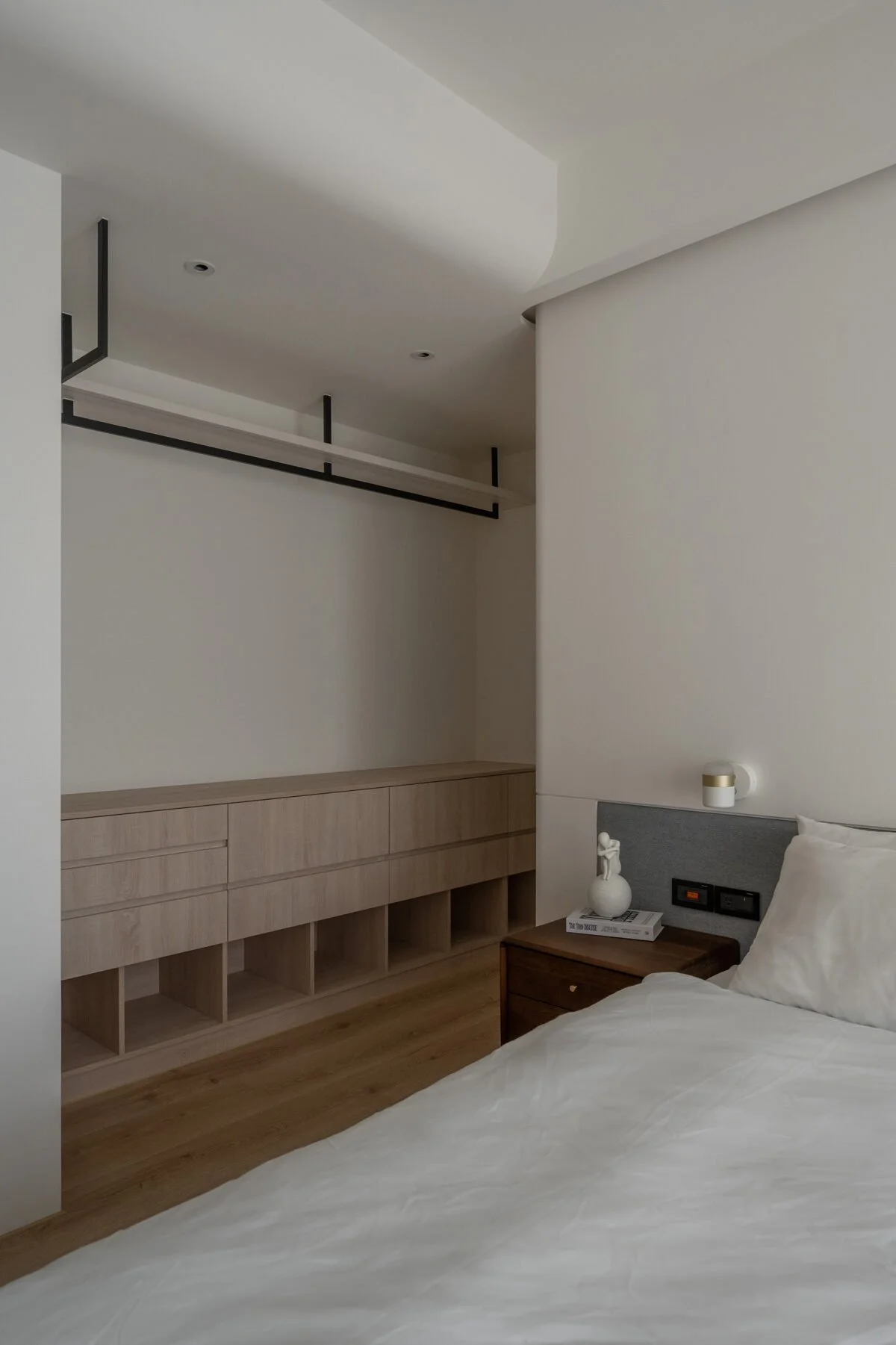 A minimalist hotel room corner with a bed and nightstand, decorated with a white vase and a small sculpture, wooden cabinet, and a wall-mounted lamp.