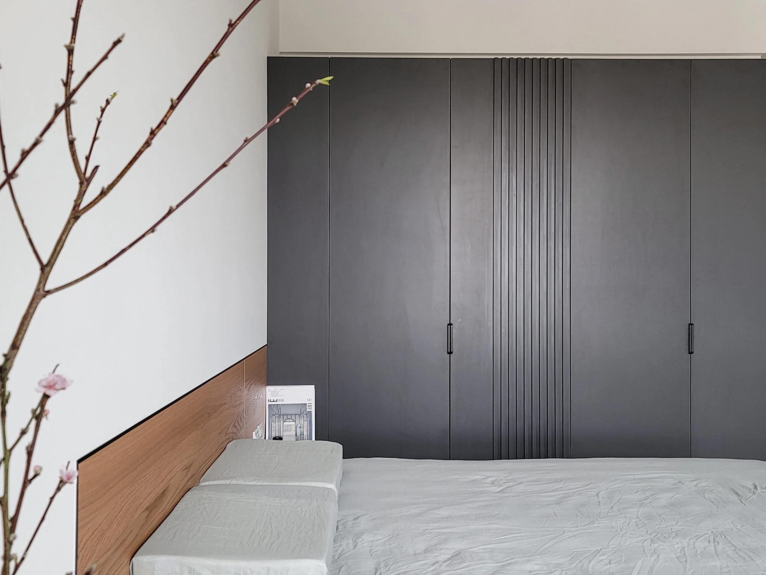 Interior of a modern room with a wooden ledge, gray wardrobe with black handles, and a white wall. A magazine or catalog is on the ledge, and pink blossoms are in the foreground.