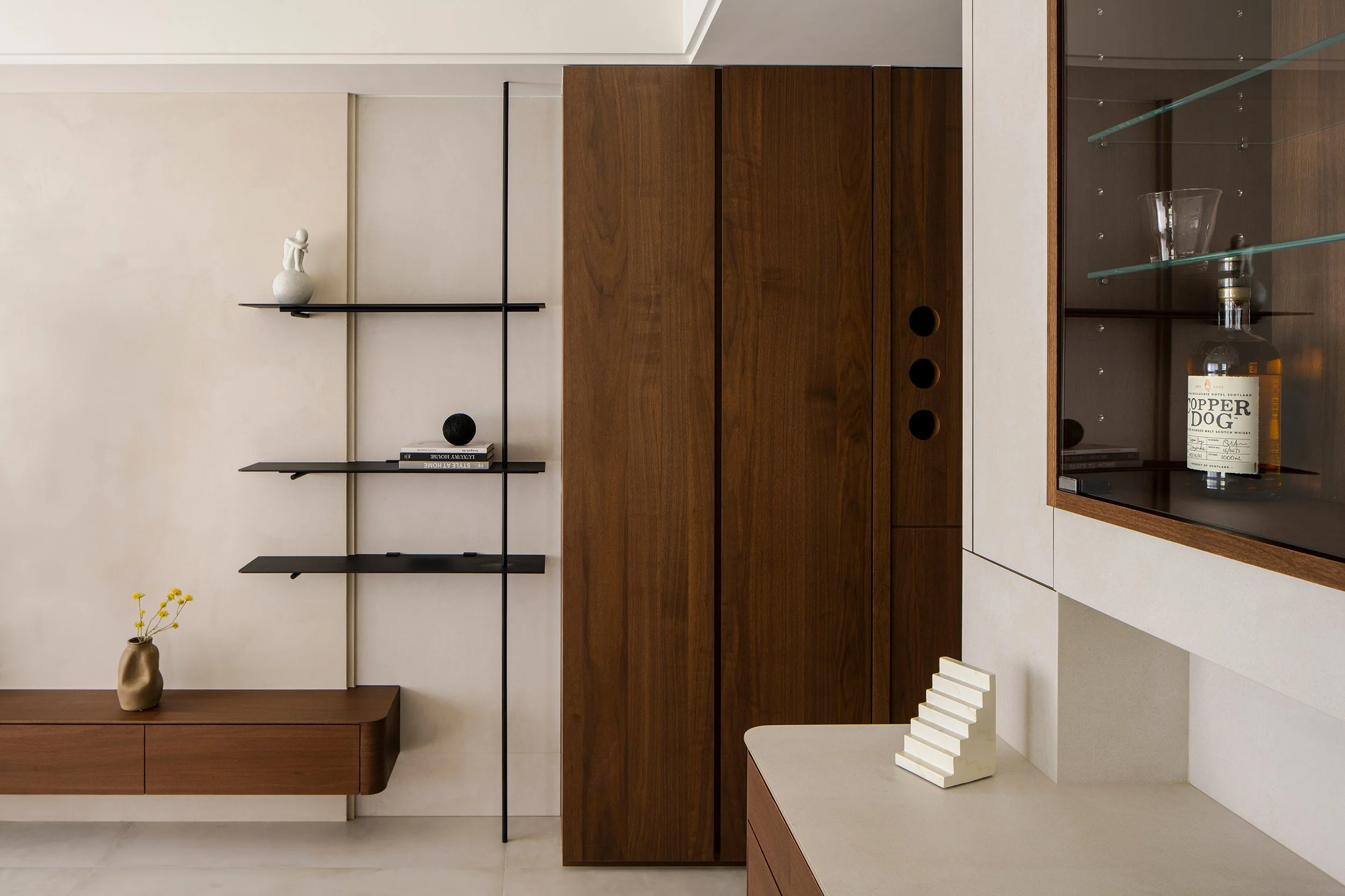 Interior view of a modern living space with minimalist decor. Features a white wall with black floating shelves holding decorative items, including a white sculpture and books. A tall beige vase with yellow flowers stands on a dark wooden bench. To the right, wooden cabinets with dark circular openings and a glass display case containing a bottle of whiskey and a glass are visible.