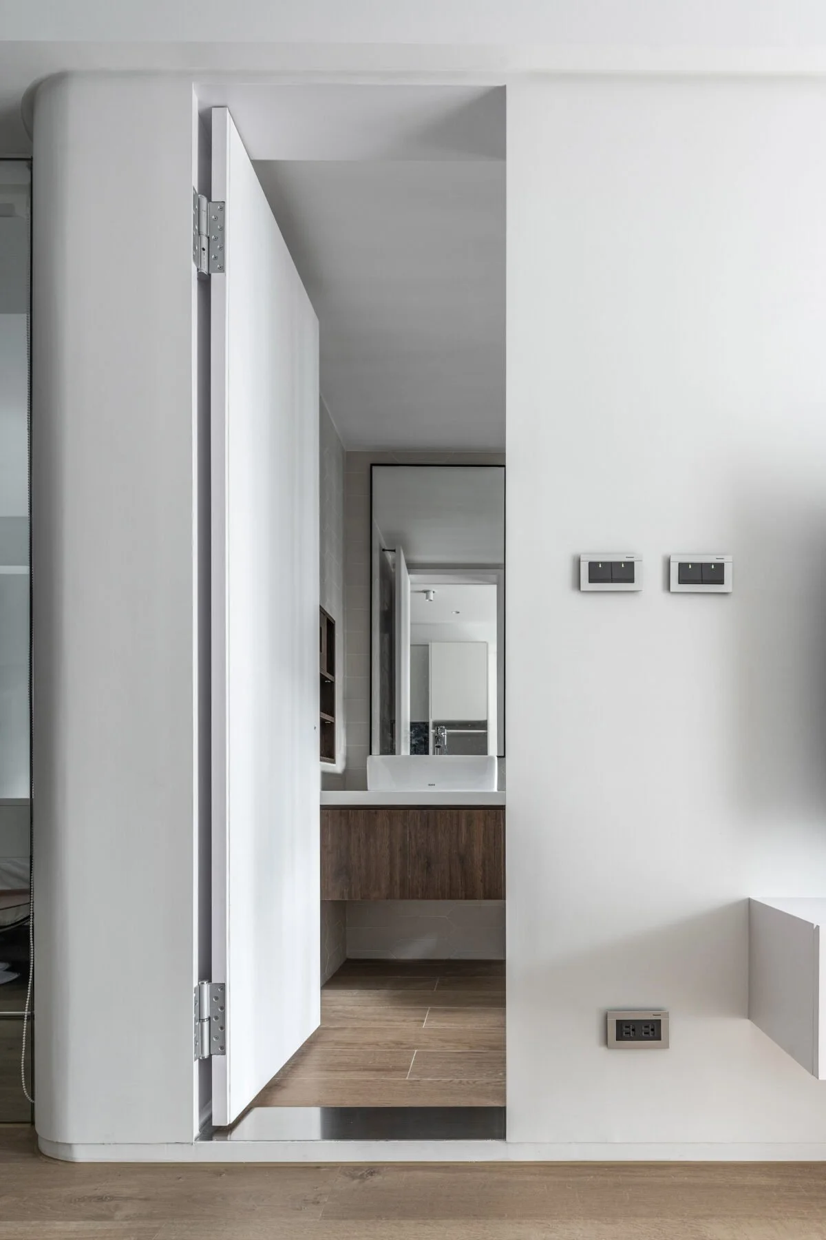 Interior view of a modern bathroom and hallway with white walls, a wooden vanity with a sink, a large mirror, and two black light switches. The hallway features a white sliding door and a floor with wooden planks.