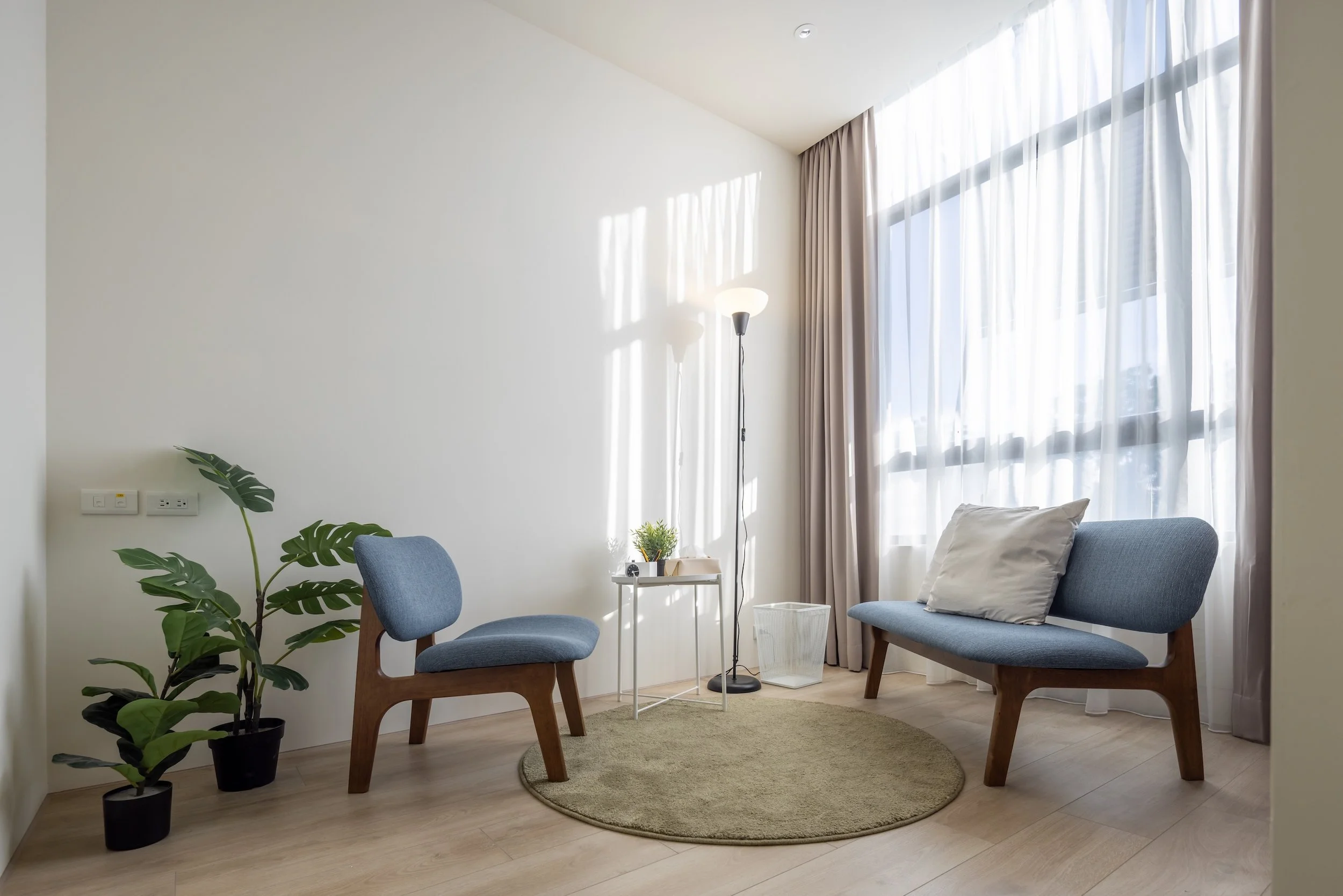 A modern living room with white walls, large windows with sheer curtains, two blue upholstered chairs, a white side table with plants, a floor lamp, and a pillow on one of the chairs.
