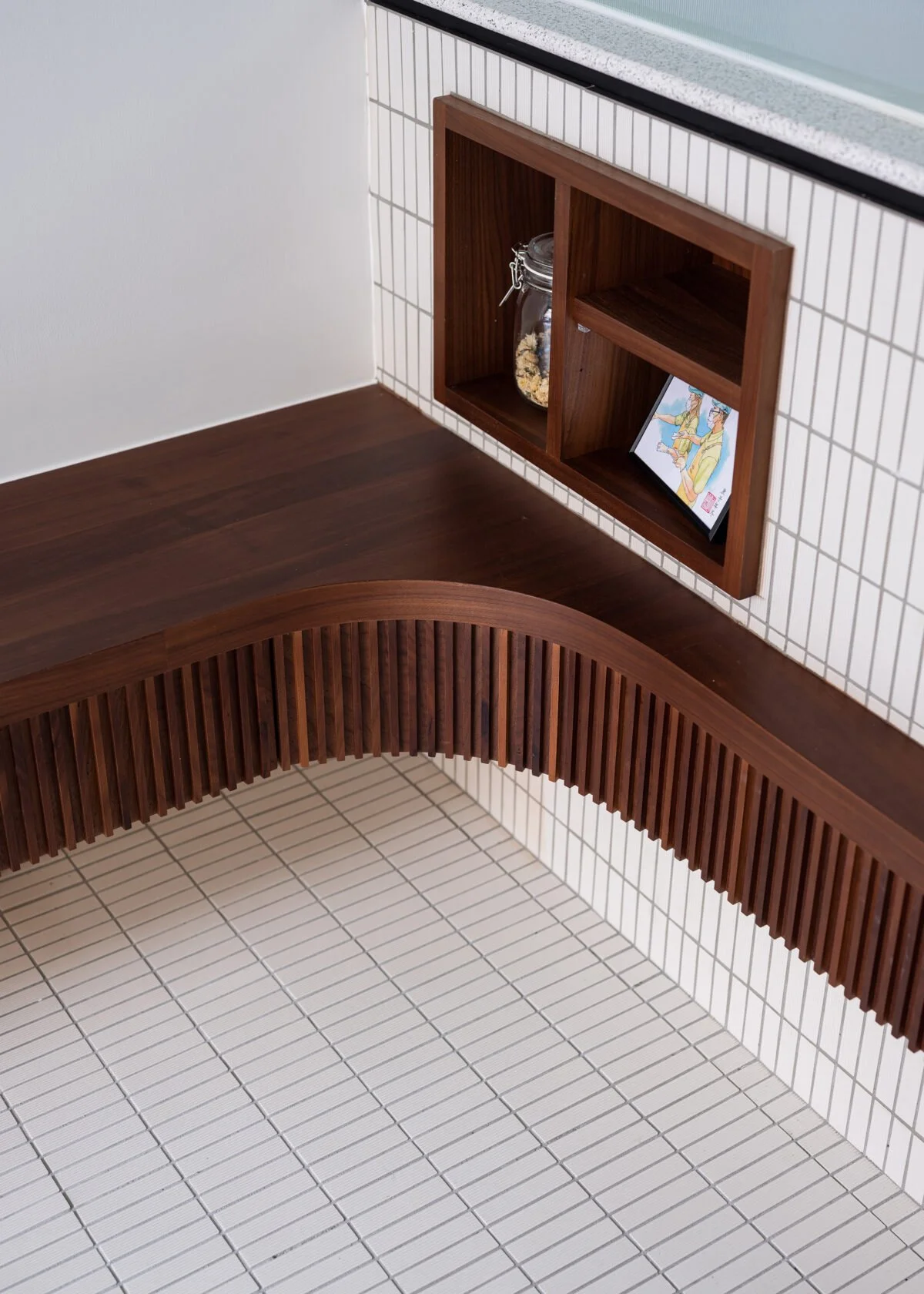 Close-up of a corner with a white tiled wall, a dark brown wooden shelf with a jar of cookies and a framed picture, and a curved wooden slat decoration on the white tiled floor.