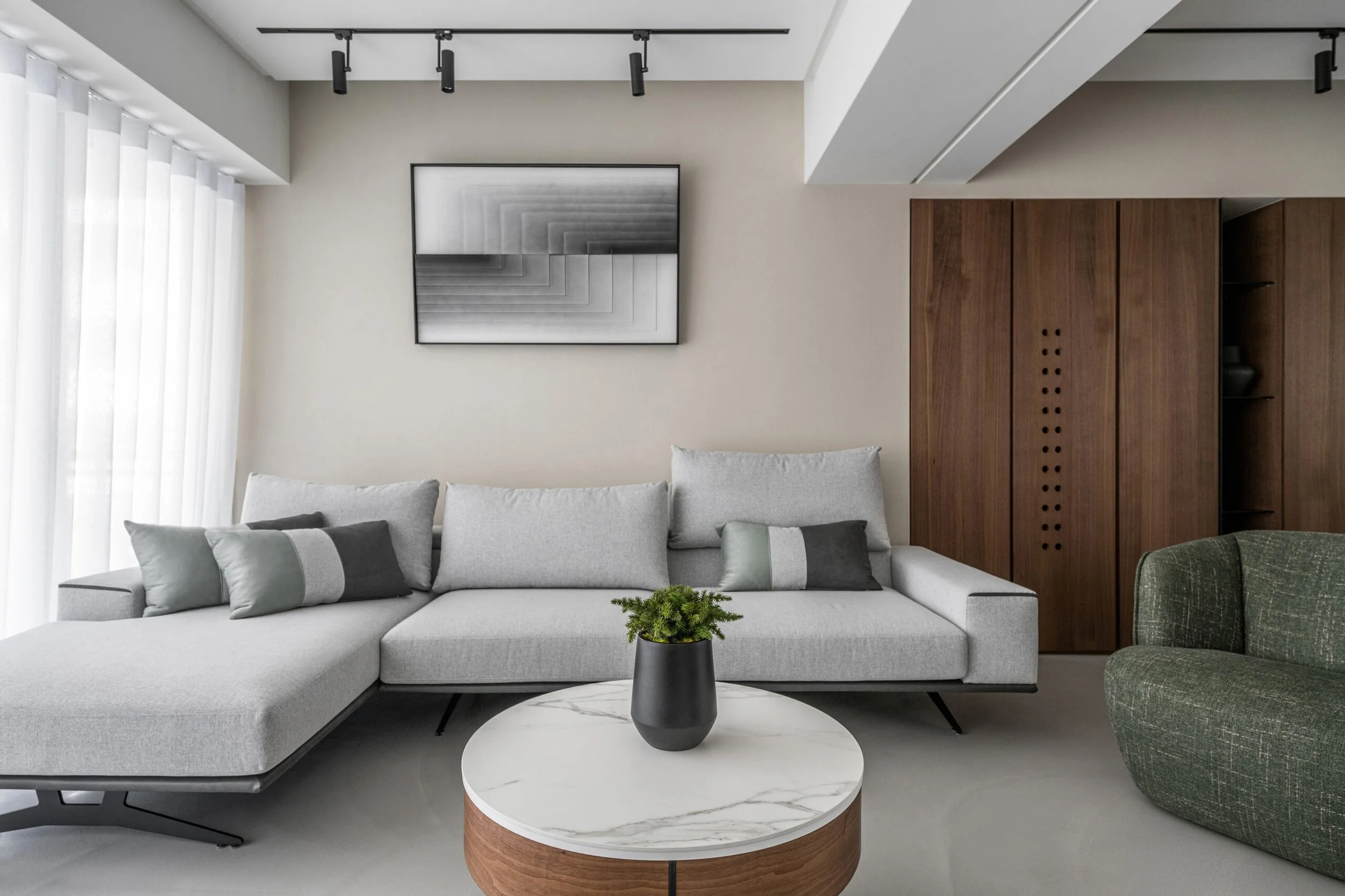 Modern living room with white sectional sofa, gray pillows, round marble-top coffee table, black vase with green plant, abstract artwork on beige wall, wooden cabinet, and black ceiling track lights.