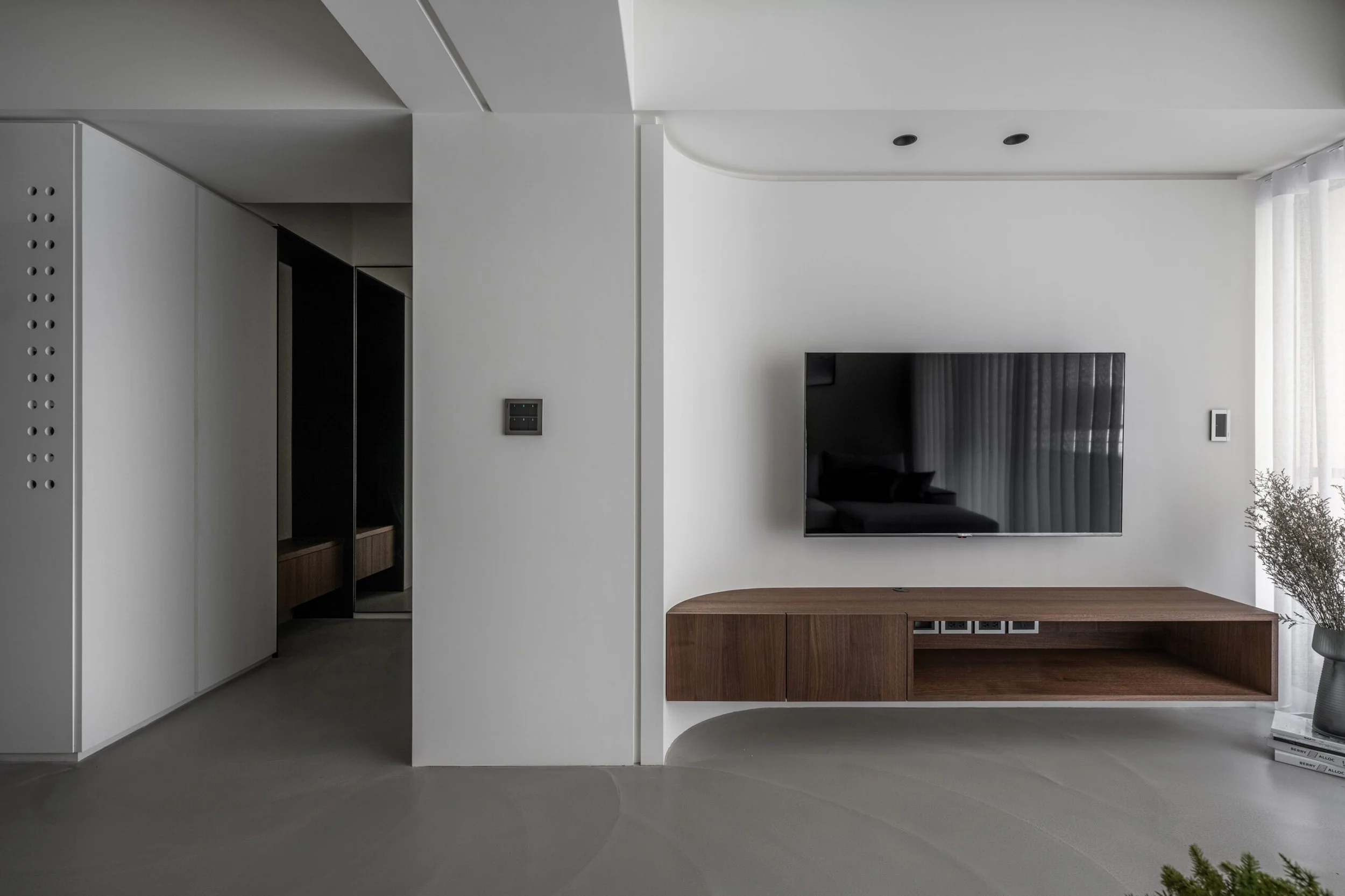 A modern minimalist living room with a large flat-screen TV mounted on a white wall, beneath a wooden storage cabinet, with a small plant to the right and a floor-to-ceiling window with curtains on the far right.