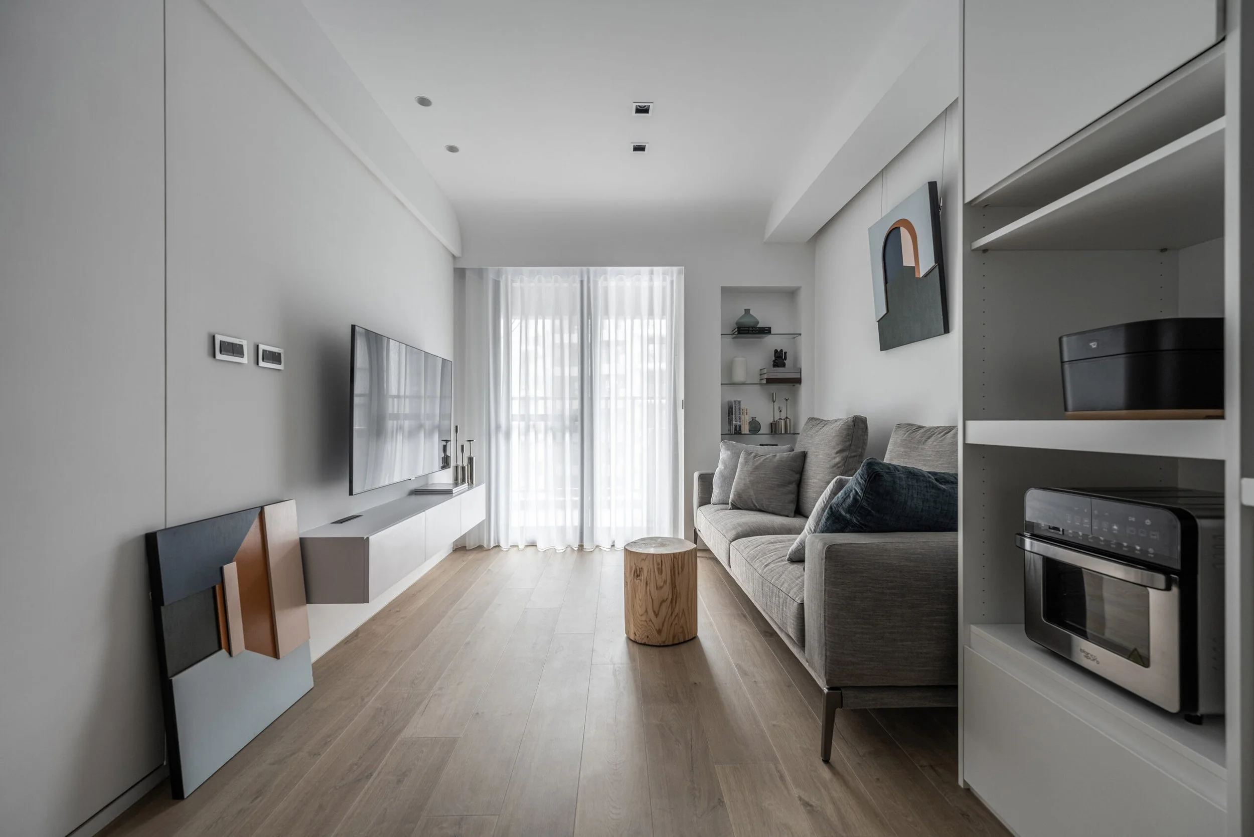 Interior view of a modern living room with white walls, a grey sofa, a small wooden side table, a TV mounted on the wall, bookshelf with decorative items, and large windows with white curtains.