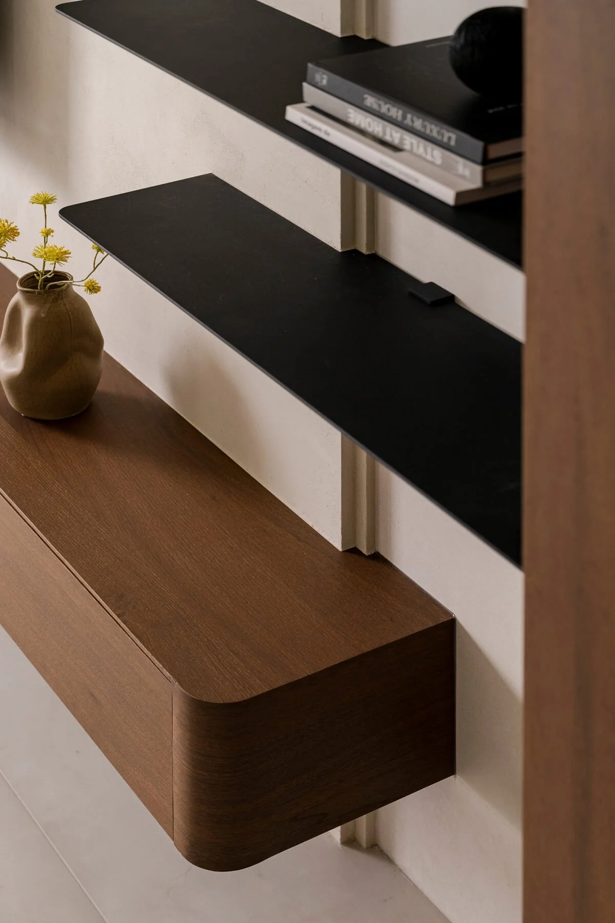 A wall-mounted minimalist shelving unit with black and wood finishes, decorated with books and a small ceramic vase with yellow flowers.