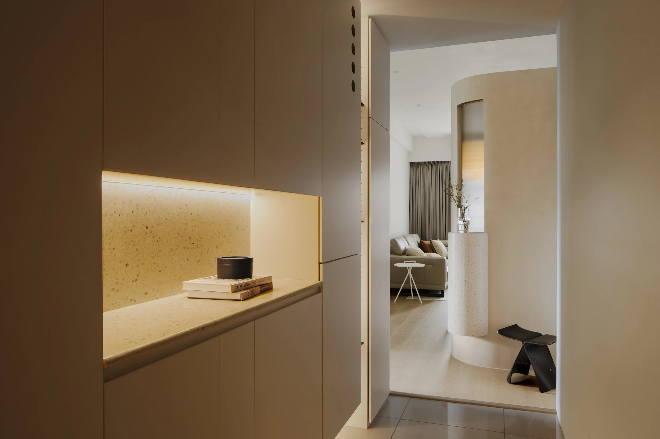 Modern minimalist interior space with beige and white tones, featuring a marble countertop with a black bowl and stacked books, leading to a living room area with a beige sofa, a small white side table, and tall window with curtains.