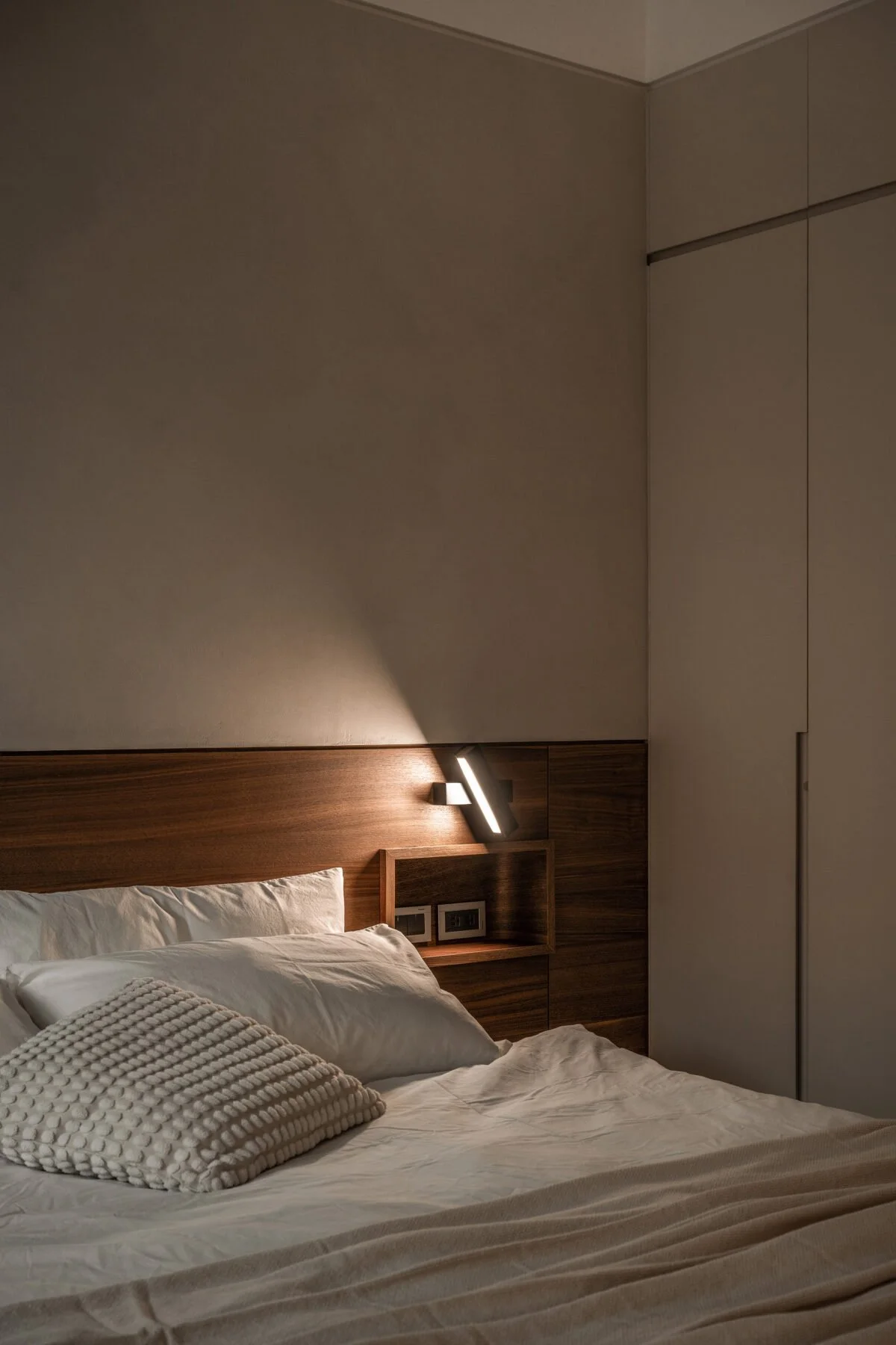 A cozy hotel bedroom with a bed, white bedding, and decorative pillows, illuminated by a modern wall lamp mounted on a wooden headboard.