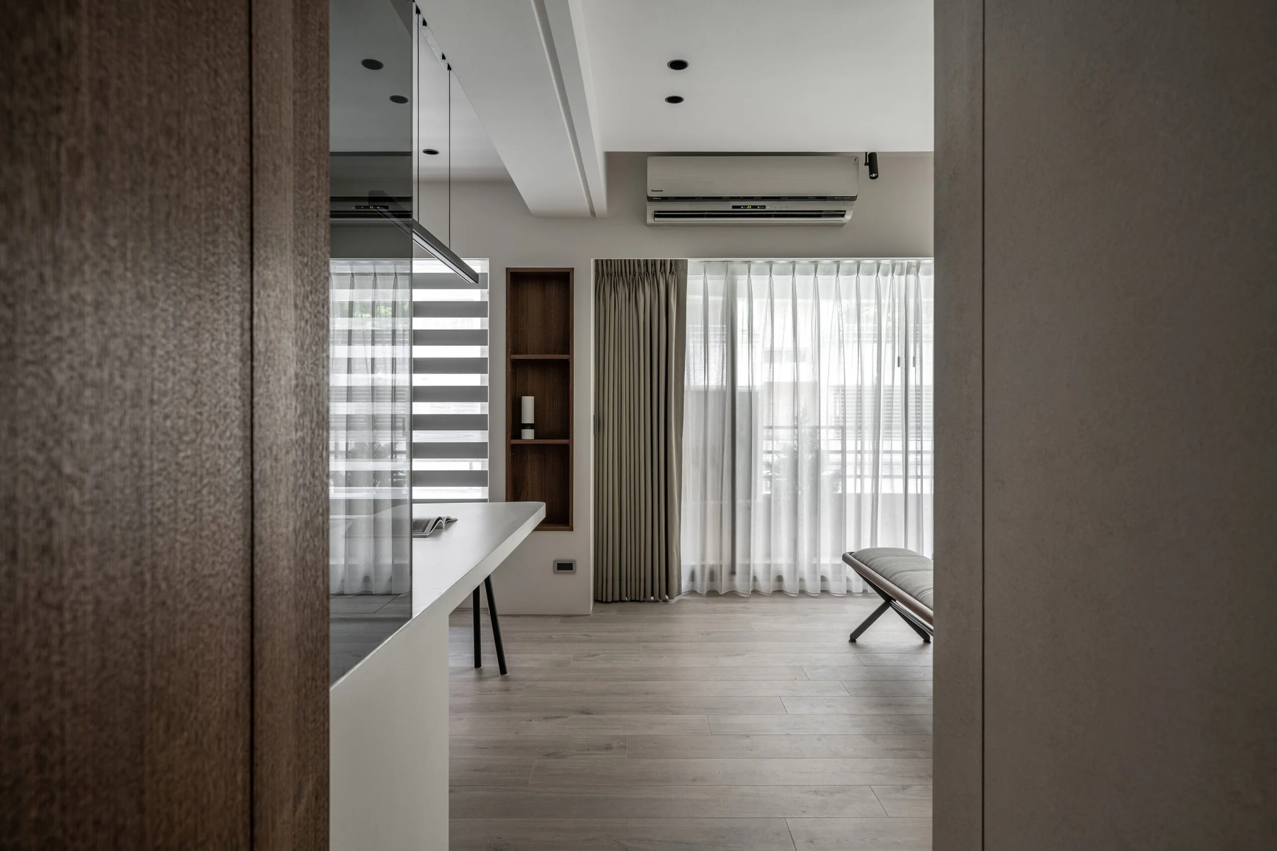 Modern living room with large windows, beige curtains, a wall-mounted air conditioner, a bench, and a small wooden shelving unit.