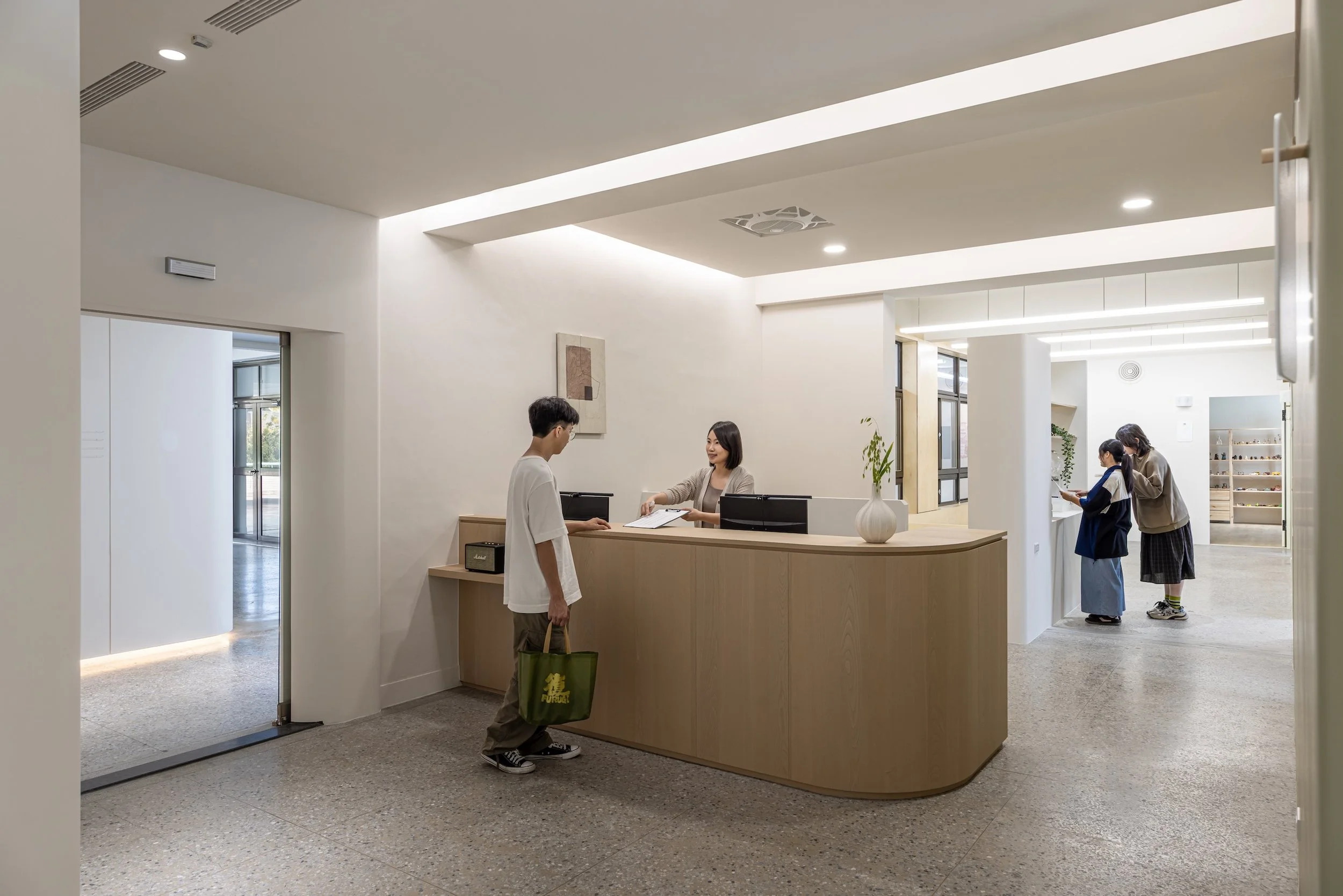 Interior of a modern reception area with a wooden curved reception desk, a woman staff member behind it, and two women on the right side engaging with the staff. A man with a green tote bag is standing near the reception. The space has white walls, a picture hanging on the wall, large windows, and ceiling lighting.