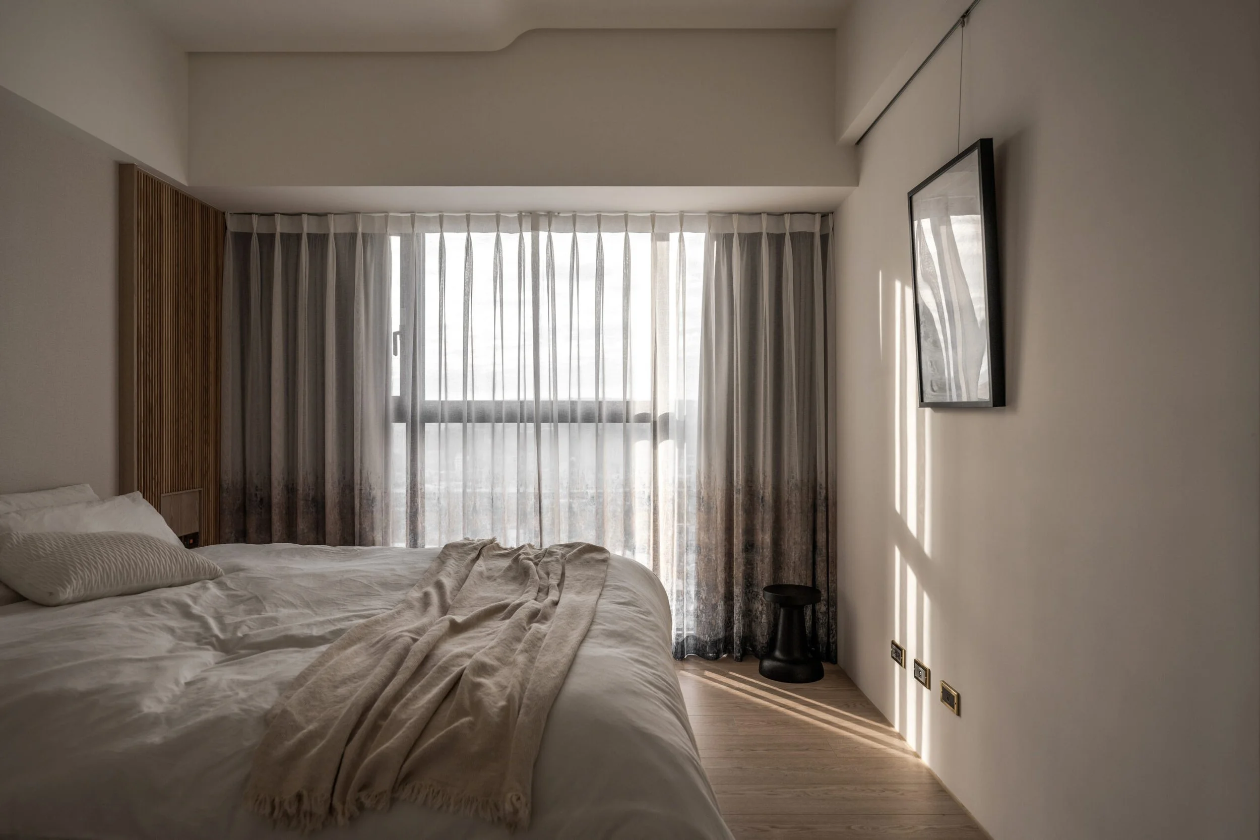A bedroom with a large window covered by sheer curtains, a bed with white bedding and pillows, a black side table, dark curtains, and a framed picture on the wall.