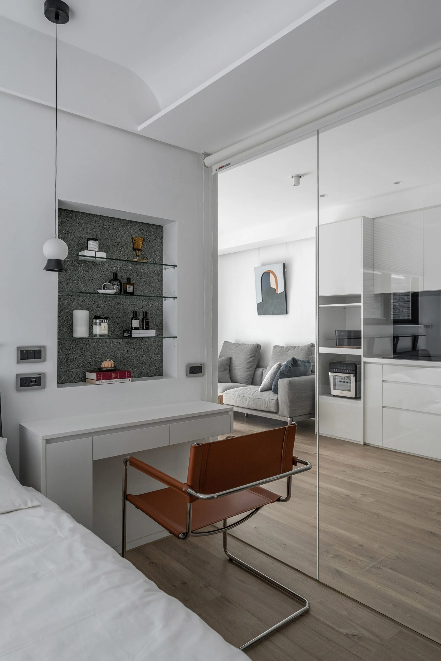 Modern living room with a gray sofa, abstract artwork on white wall, glass shelves with decorative items, and a glass wall with view into a kitchen space.