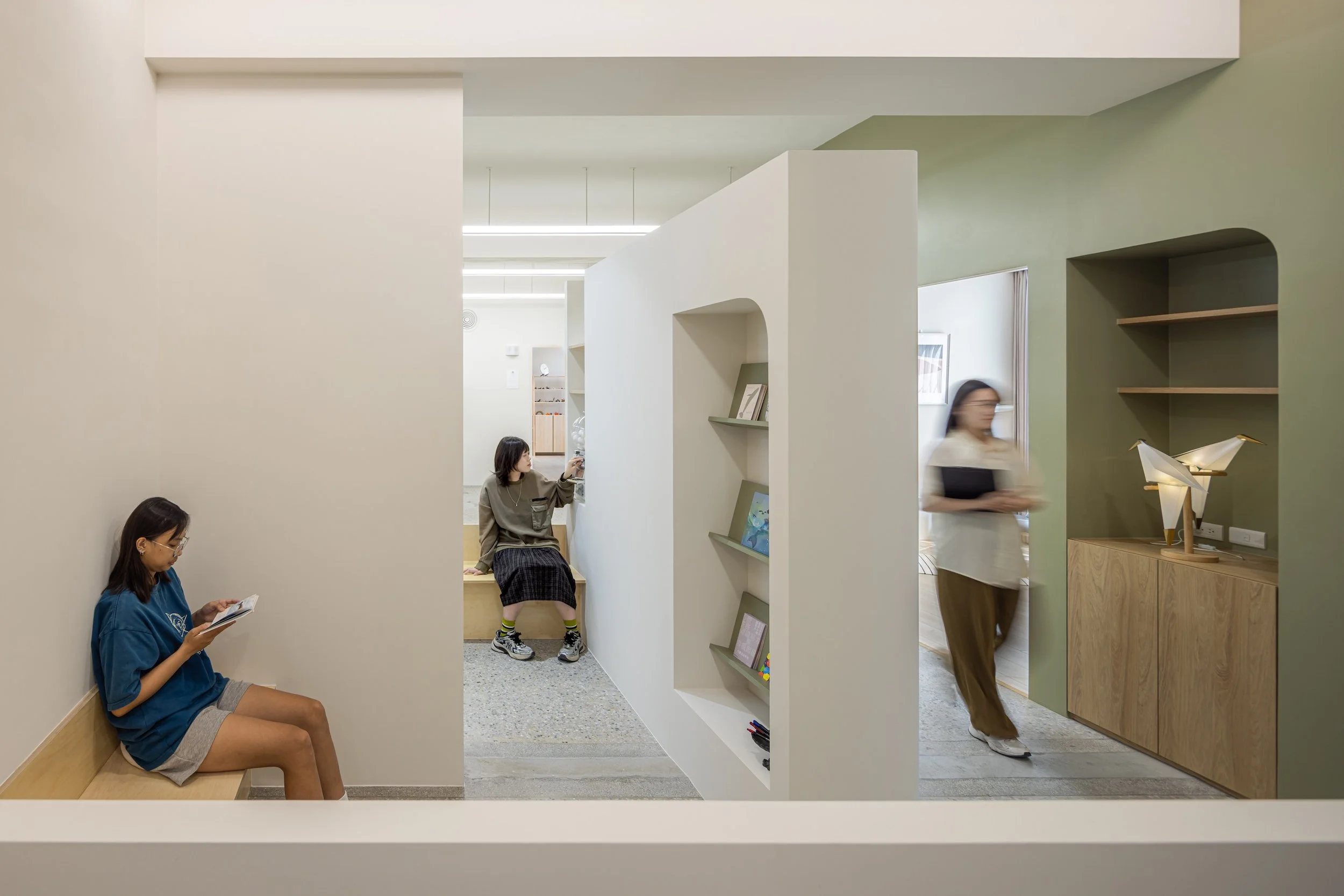 Interior of a modern, minimalist space with people engaging in different activities, including sitting, walking, and reading, separated by partition walls and shelves.