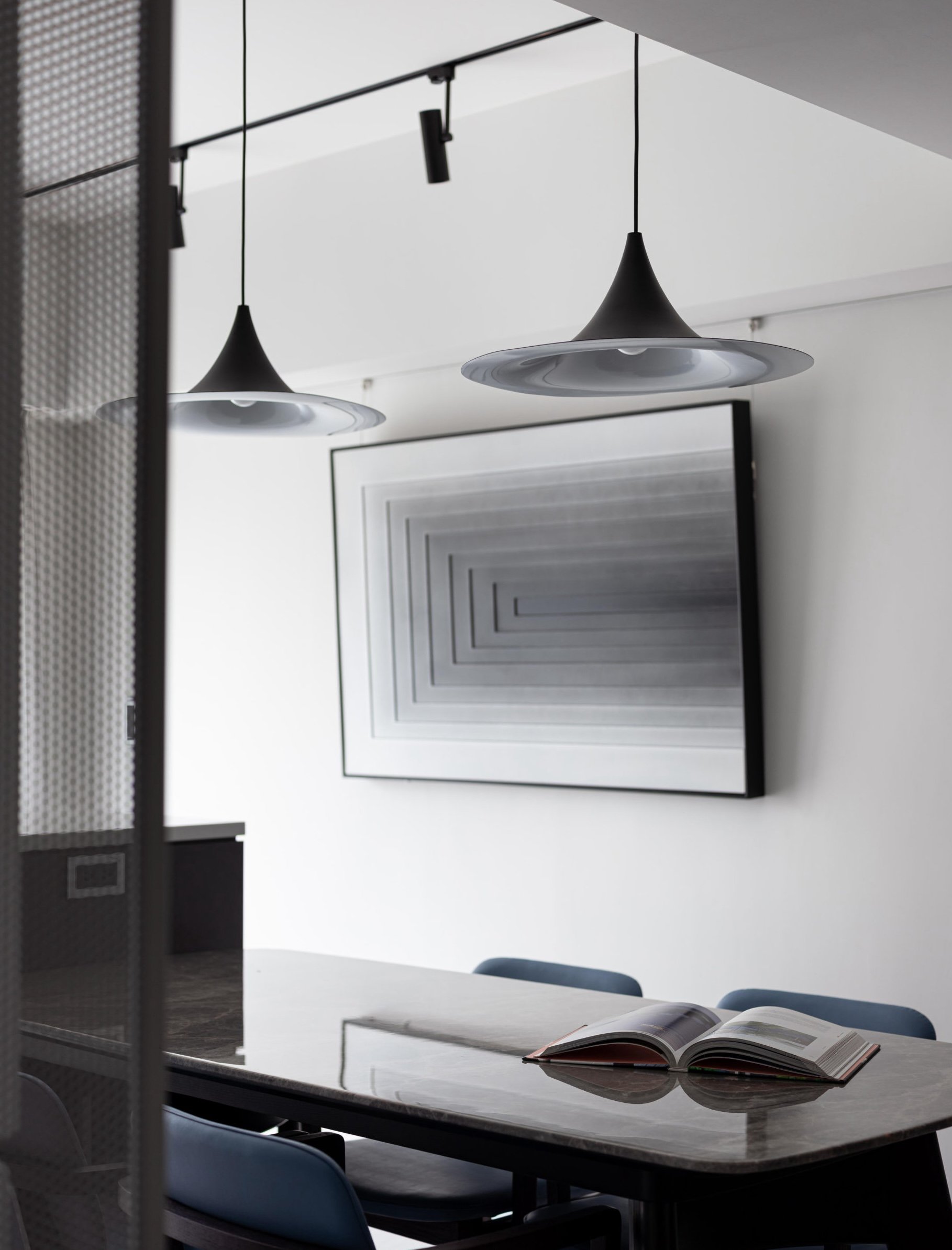 A modern office conference room with two black pendant lights, a large framed geometric artwork on the white wall, a white table with an open magazine, and blue chairs.