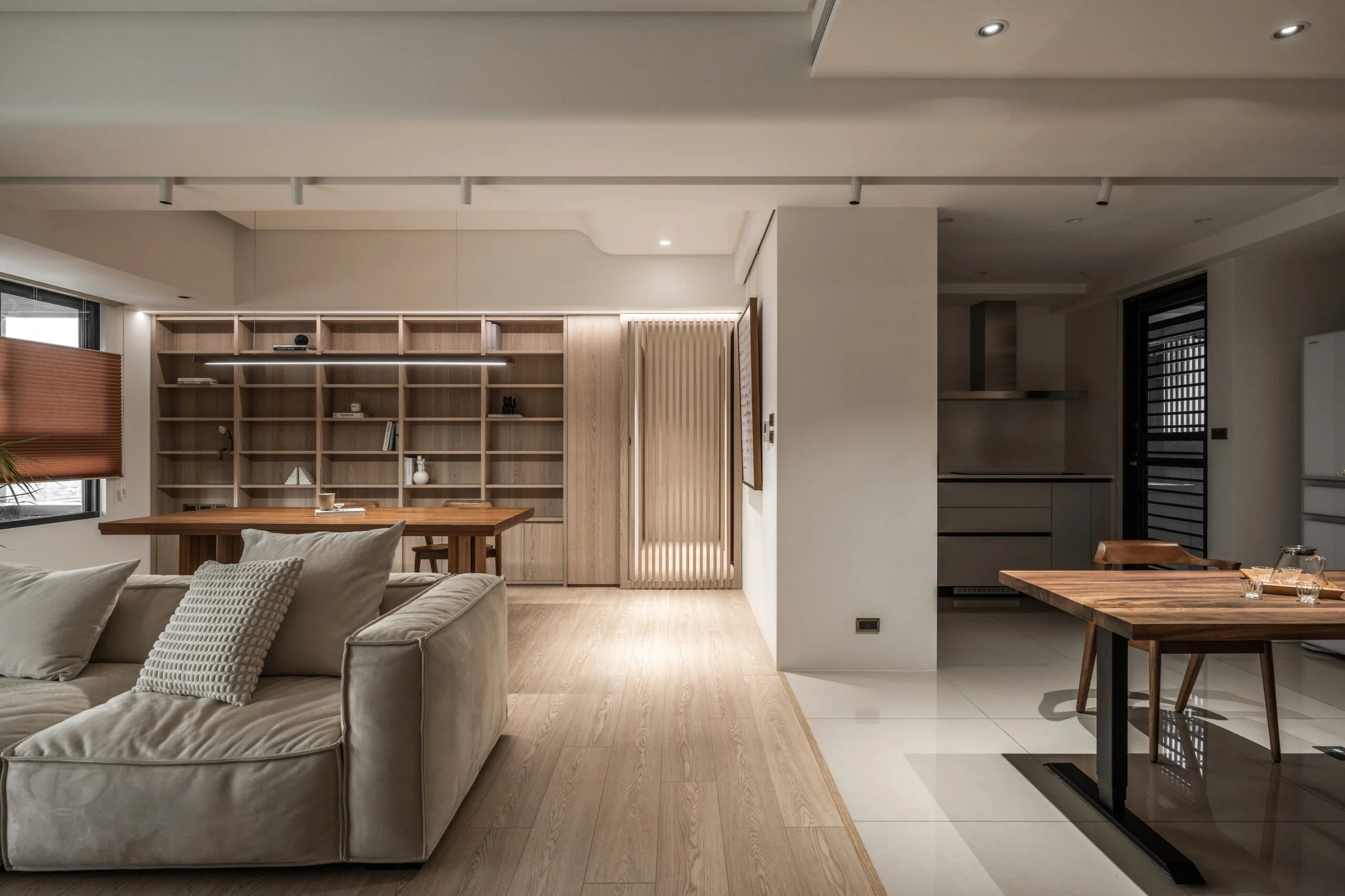 Open-concept living room and kitchen with wooden floors, built-in wooden bookshelf, beige sofa with pillows, wooden dining table, and modern lighting fixtures.