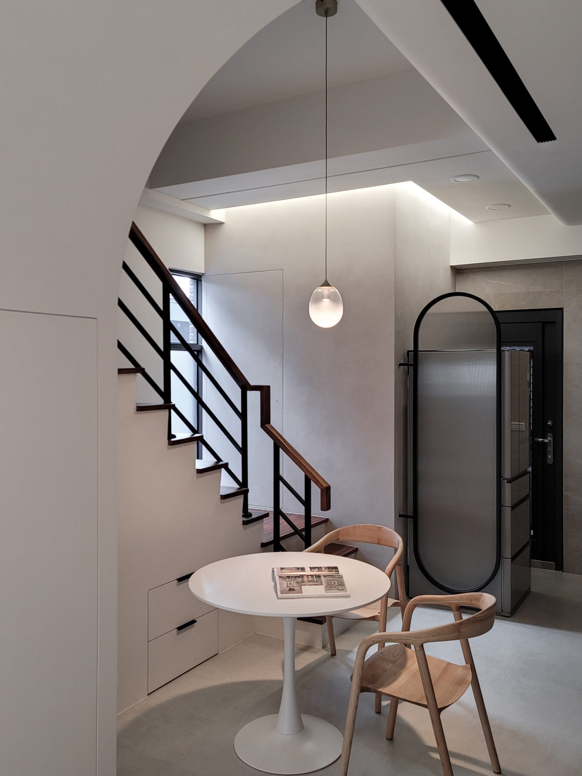 Modern interior with a staircase, hanging pendant light, white round table with magazines, and wooden chairs in a bright room.