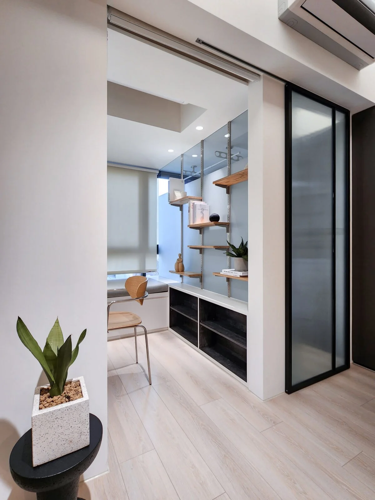 Modern, minimalist office corner with beige walls, wooden flooring, a desk with a wooden chair, a window with a white roller shade, and a shelving unit with books, decorative items, and a potted plant. There is also a black and glass sliding door and a wall-mounted air conditioning unit.