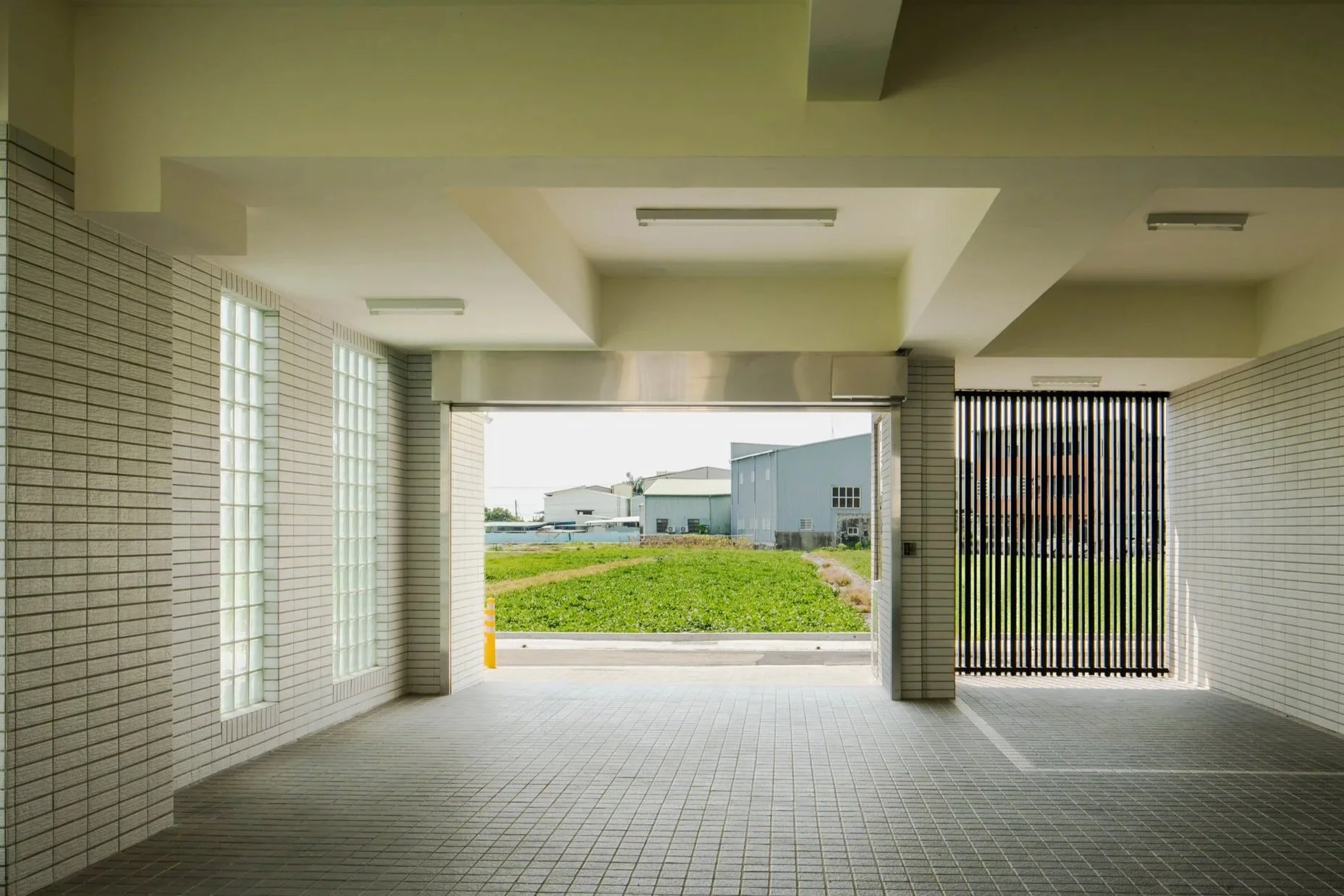 自遮蔽廊道下方往開闊處望去，視野穿過開放空間，遠處為草坪與工業廠房景致。