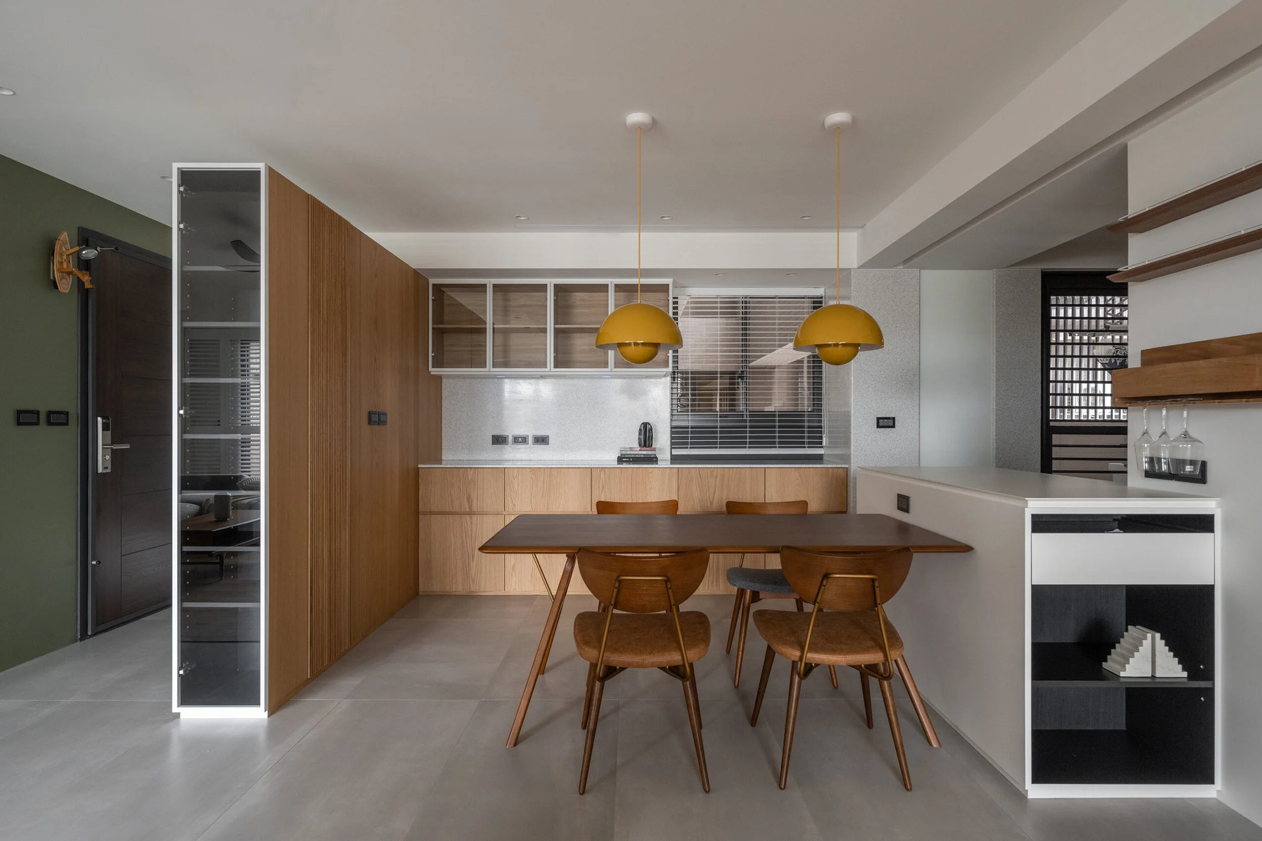 Modern kitchen and dining area with wooden furniture, yellow pendant lights, and built-in shelving with glassware.