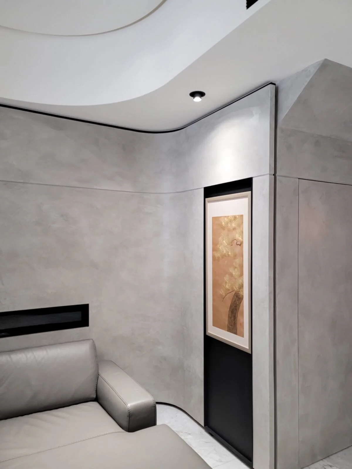 Modern living room corner with a light gray leather sofa, curved textured gray wall, a framed artwork of a tree on a narrow black wall, and a ceiling with a small recessed light.