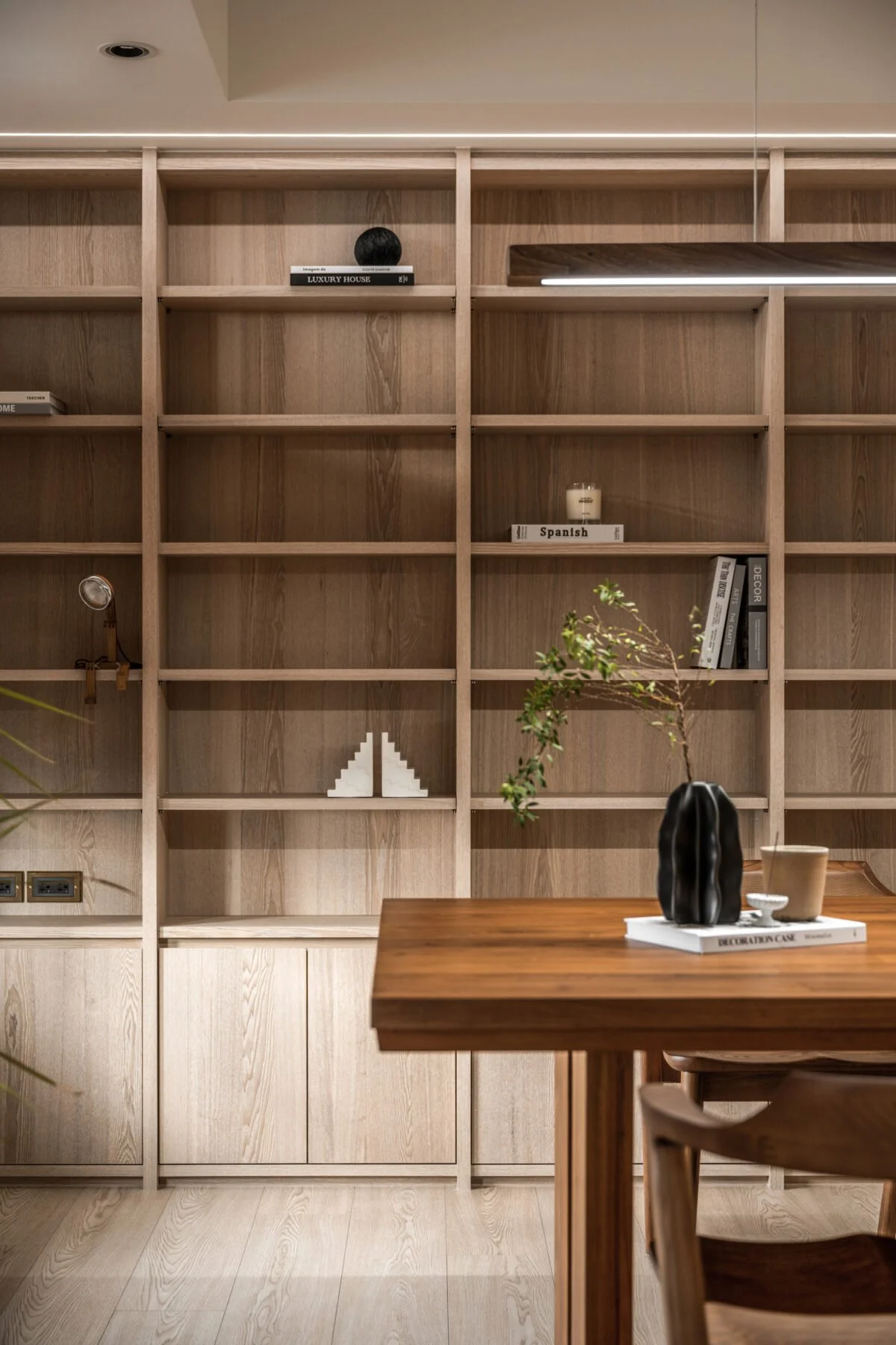Empty wooden bookshelf with a few decorative items, a potted plant on a table in front, and a chair partially visible in a modern interior space.