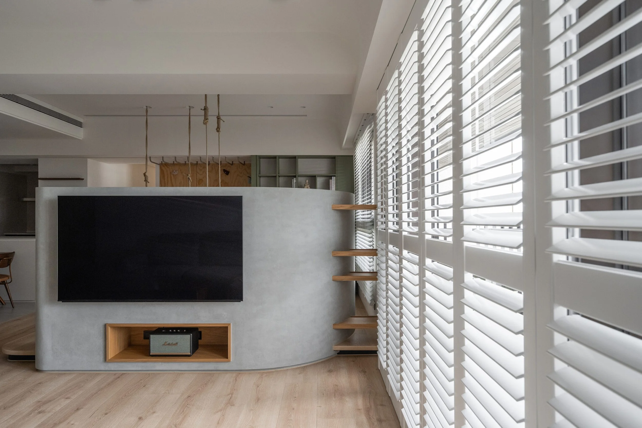 Modern living room with large white shutters on windows, a black chalkboard wall, white curved room divider with open shelving, a TV, and wooden floors.