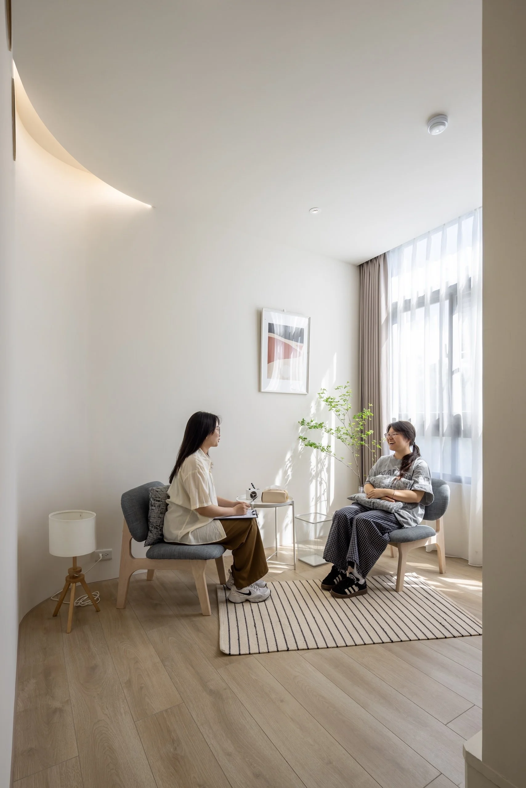 Two women sitting in a living room, engaged in conversation. One is taking notes and the other is smiling with a pillow on her lap. The room is bright with natural light, minimalist decor, a framed picture on the wall, and a striped rug on wooden flooring.