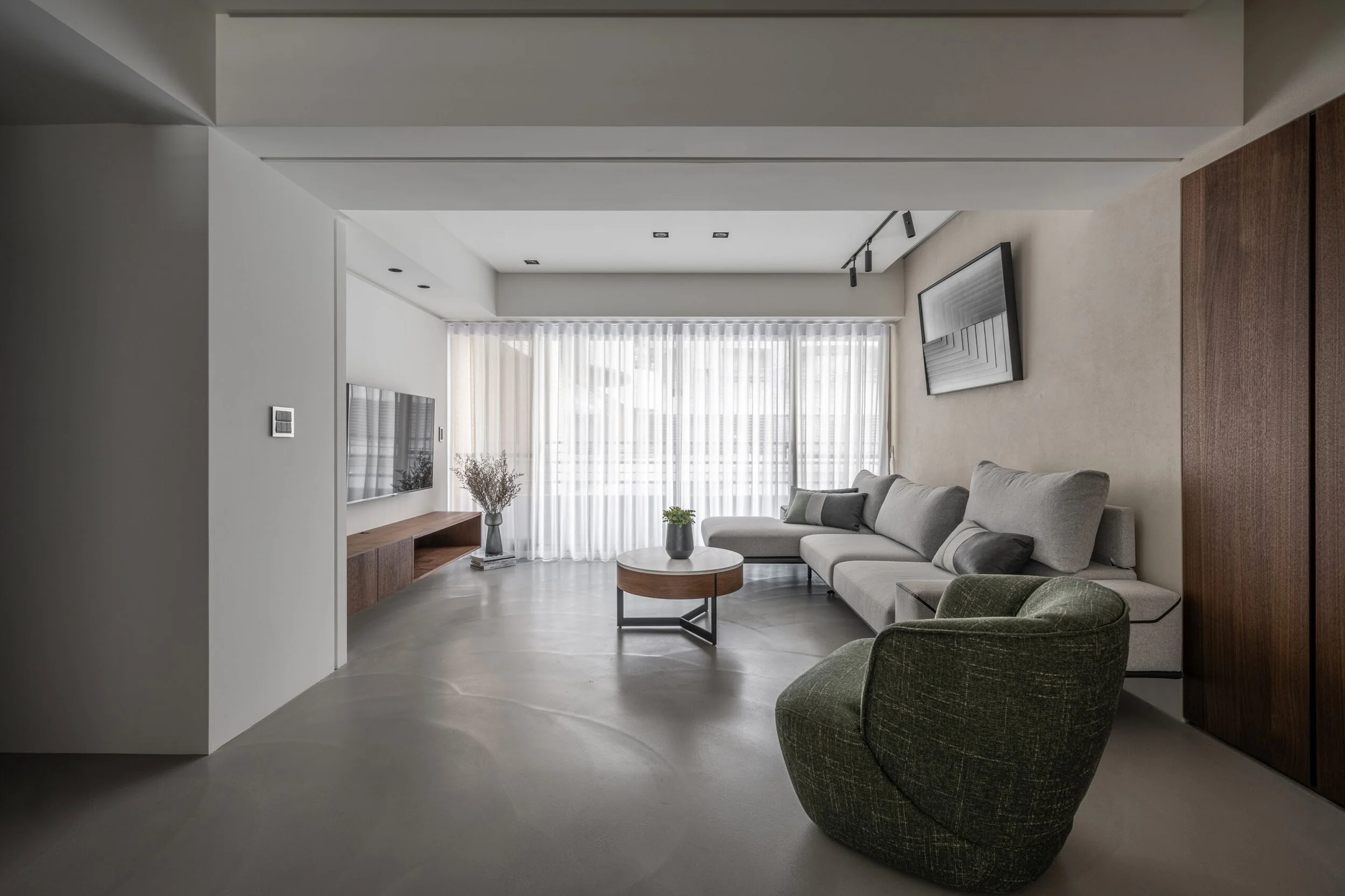 Modern living room with beige sectional sofa, green armchair, round coffee table, wall-mounted TV, large window with sheer curtains, and minimalist decor.