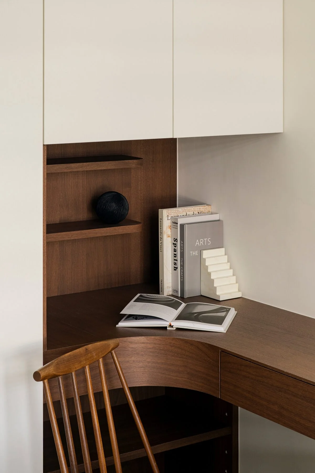 Corner of a wooden desk with a wooden chair. On the desk, there are books, one open with black and white photographs, and others upright with titles related to arts and spirituality. Shelves above the desk hold a black spherical sculpture and a small wooden item.