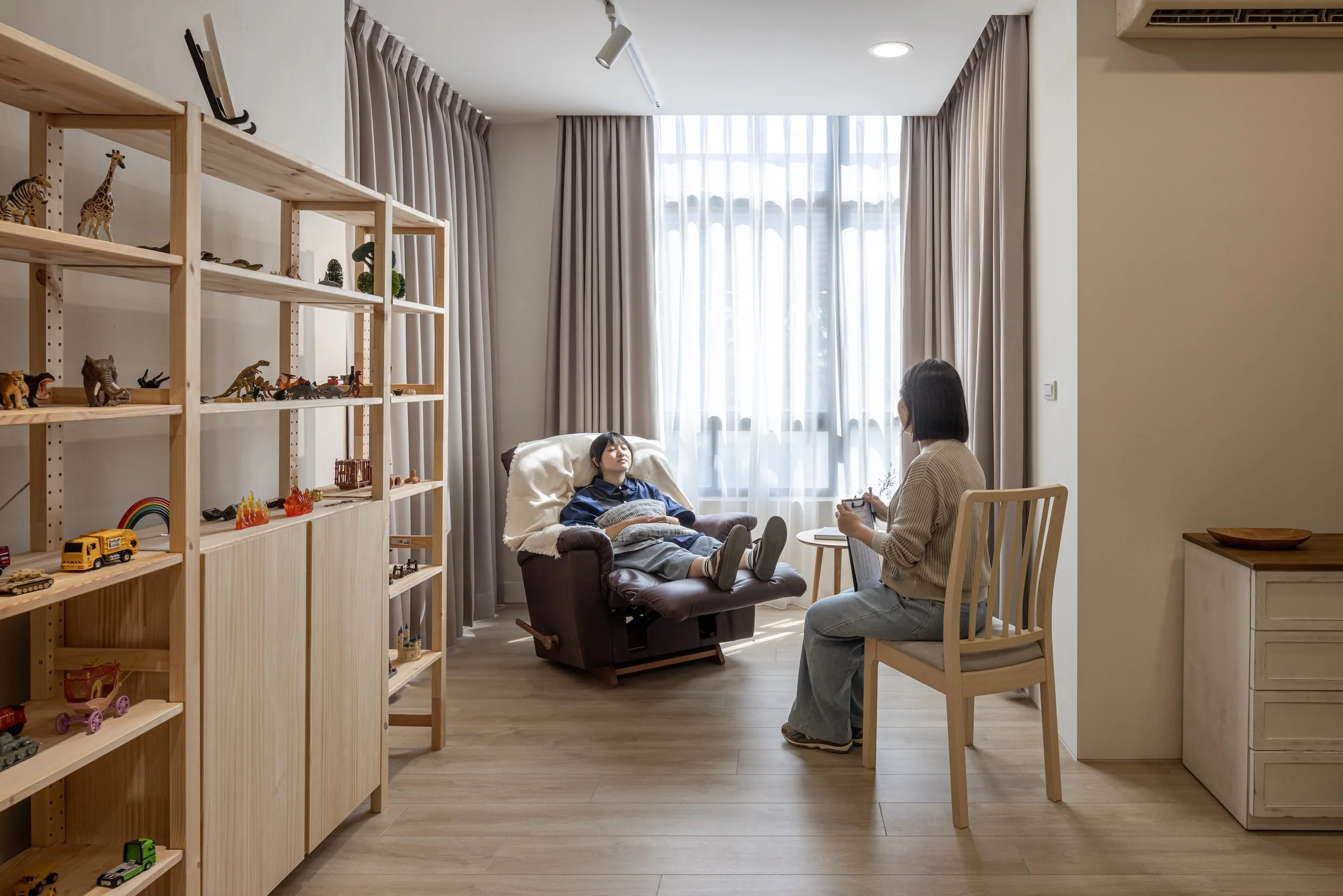 A woman is lying in a recliner chair with her eyes closed while a woman sitting on a chair next to her appears to be talking or explaining something, inside a room with large window and beige curtains. The room has wooden floors and a wooden shelf filled with toy animals and vehicles.