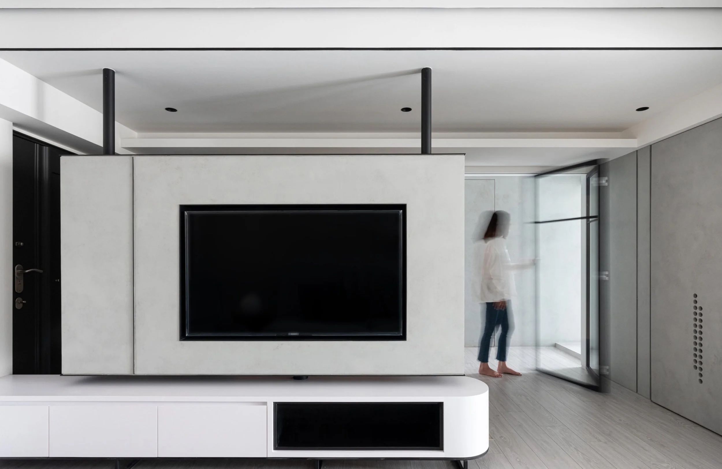 Modern living room with a large flat-screen TV embedded in a white wall-mounted unit, and a woman in a white shirt and jeans standing near the glass door on the right.