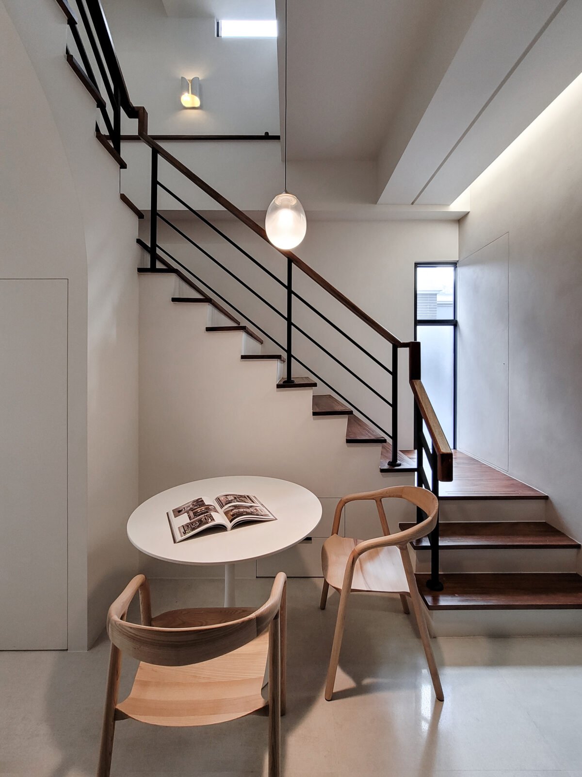 Modern minimalist interior with a staircase, wooden chairs around a small white round table, an open book, and natural light from windows.
