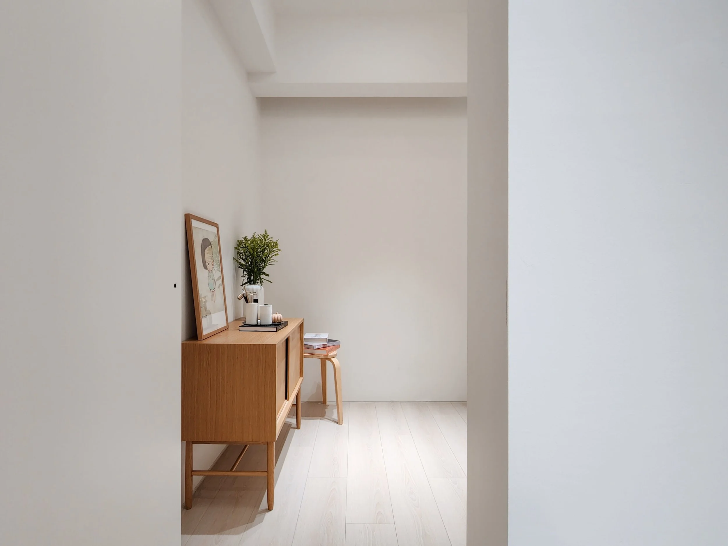 A minimalist entryway with white walls, a wooden console table holding framed art, a potted plant, candles, and a small pumpkin, and a stool underneath.