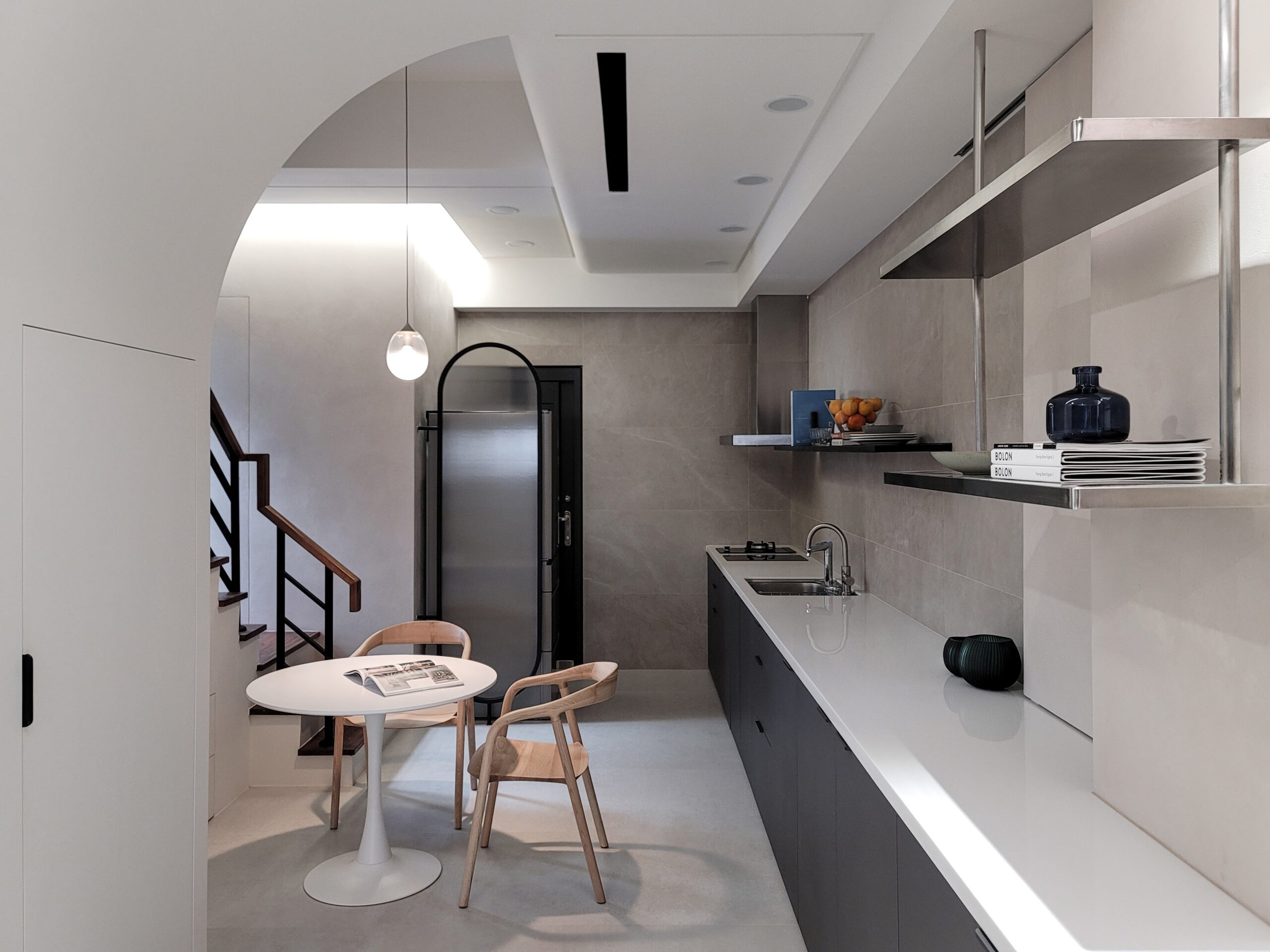 Modern kitchen with white round table and three light wood chairs, open shelving with books and decor, stainless steel appliances, pendant light, neutral tones