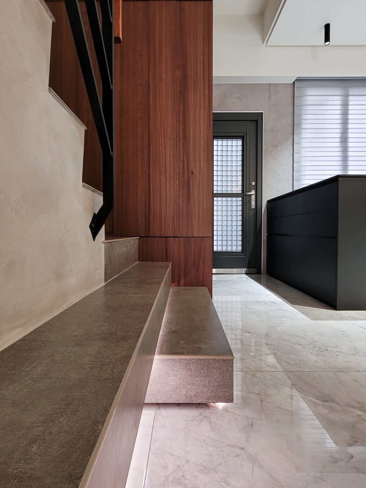 Interior view of a modern entryway with beige marble floor, a black door with glass panels, a dark gray cabinet, and a wooden wall with stairs.