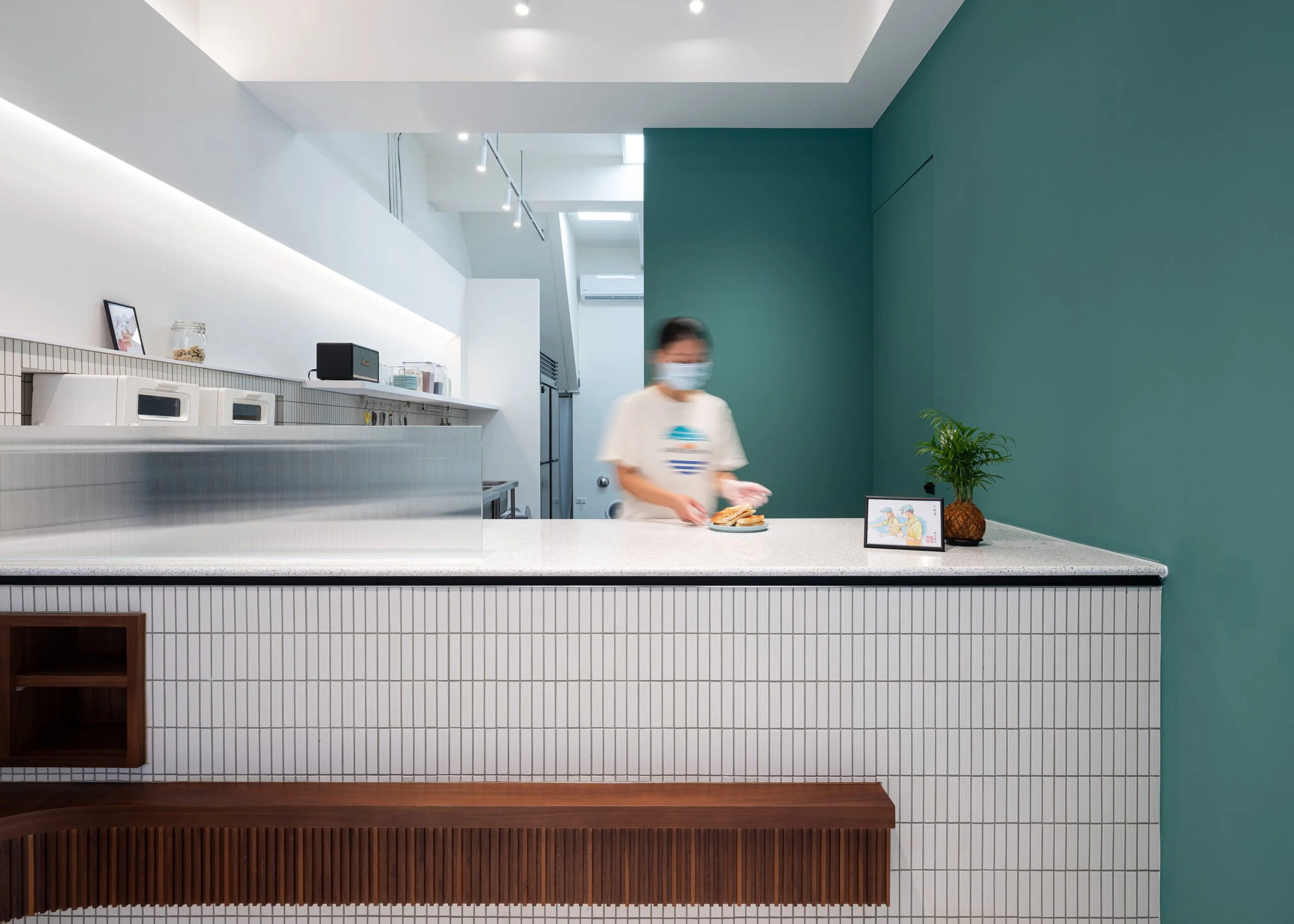 A person wearing a face mask standing behind a white counter with a plate of sandwiches, a potted plant, and framed pictures, in a modern minimalist interior with white and teal walls.
