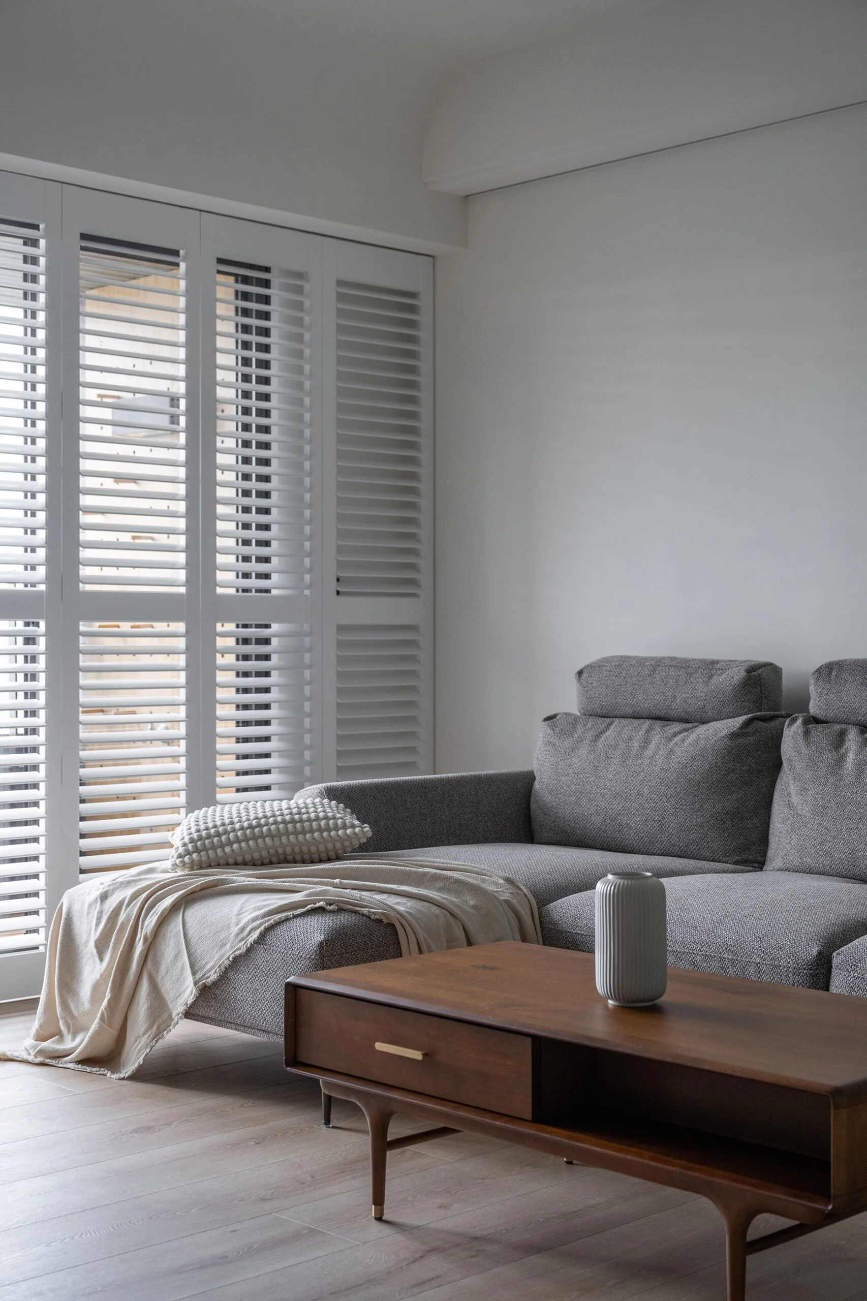 Living room corner with a grey sectional sofa, beige blanket, textured pillow, wooden coffee table with a ceramic vase, and large white window shutters.