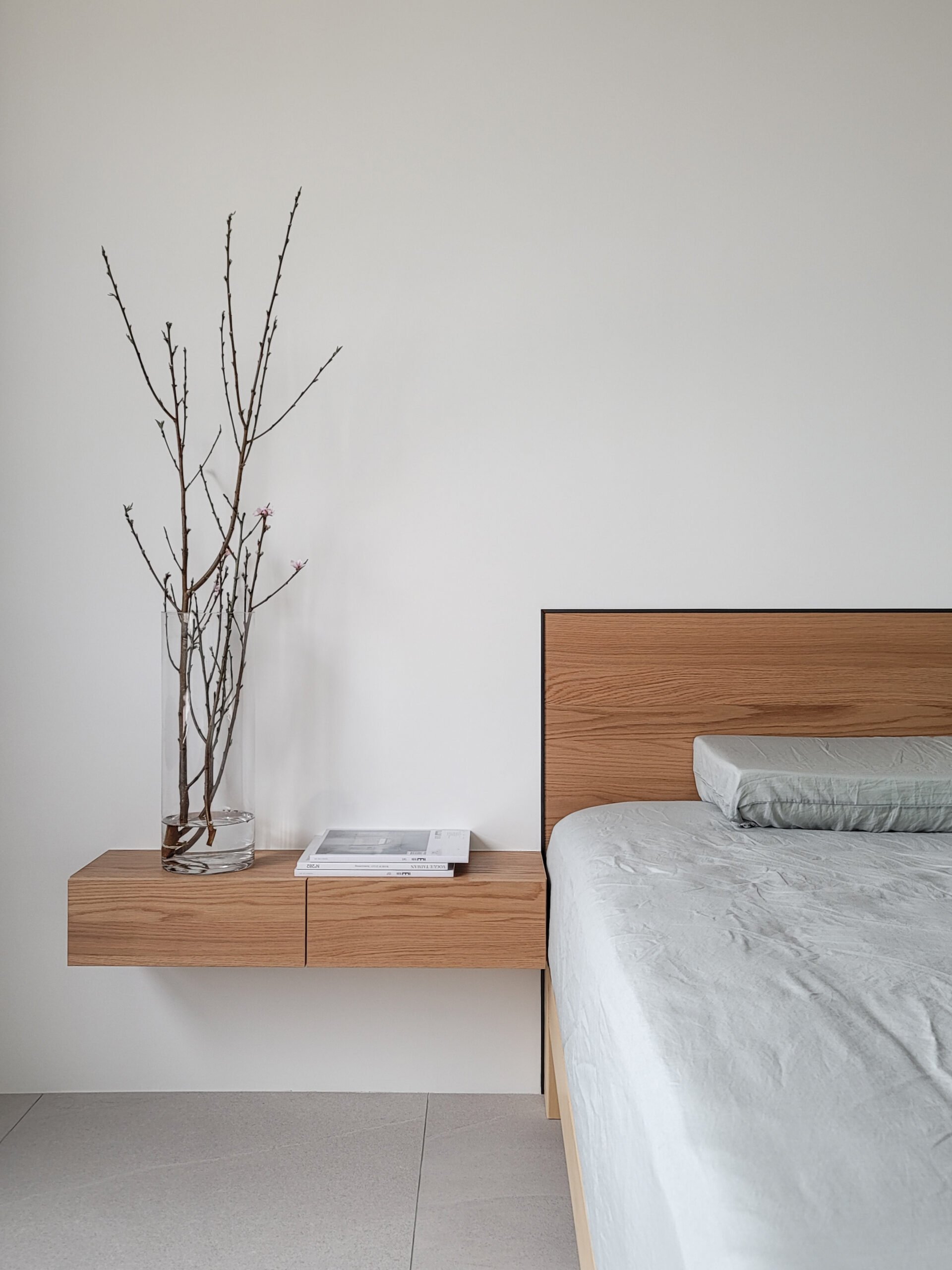 A modern bedroom corner with a white wall, a wooden nightstand with books, a glass vase with bare branches, a bed with a white comforter and a pillow, and a wooden headboard.