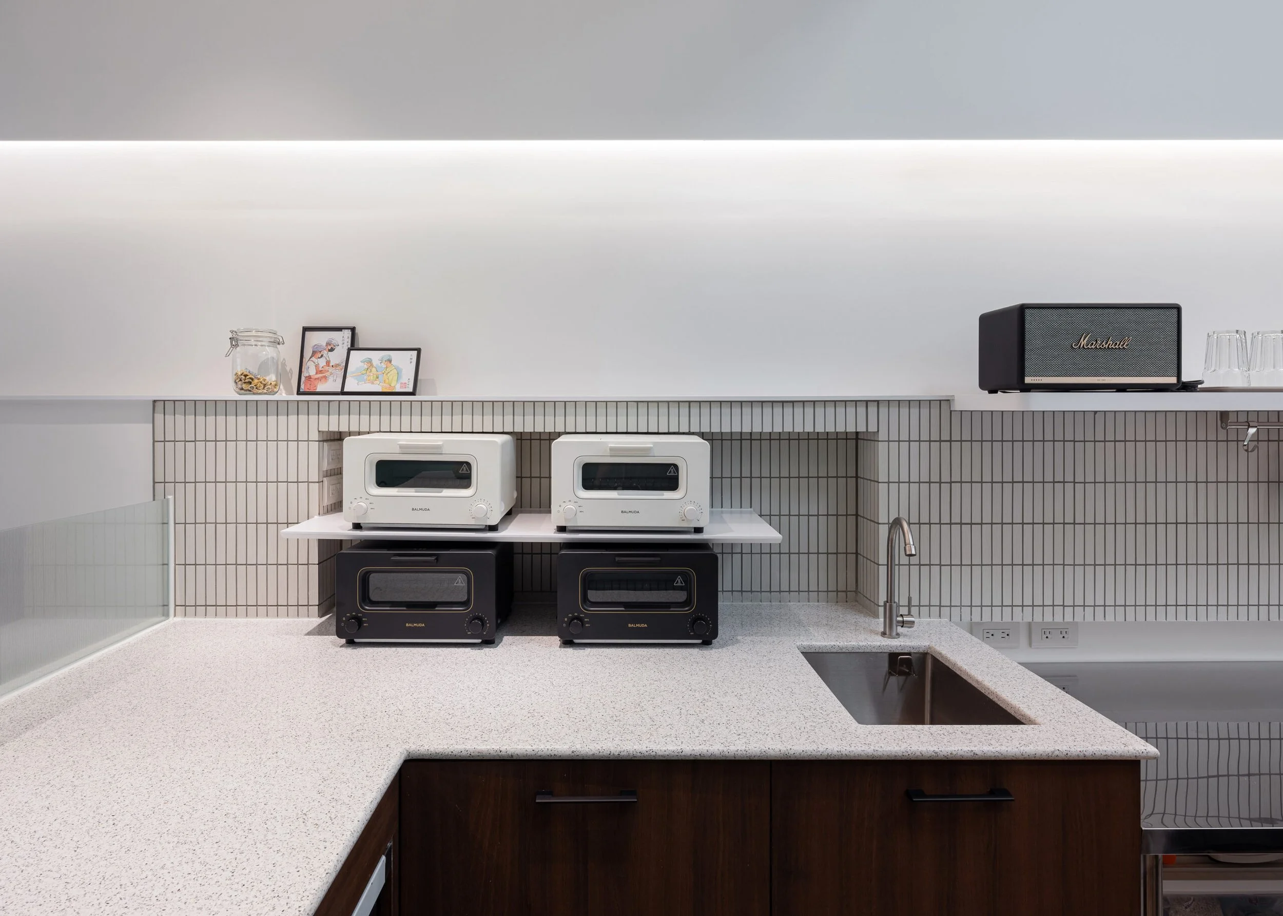 A kitchen counter with two toasters, framed pictures, a glass jar with cookies, and a Marshall speaker, with a tiled backsplash and a sink with a faucet in the foreground.